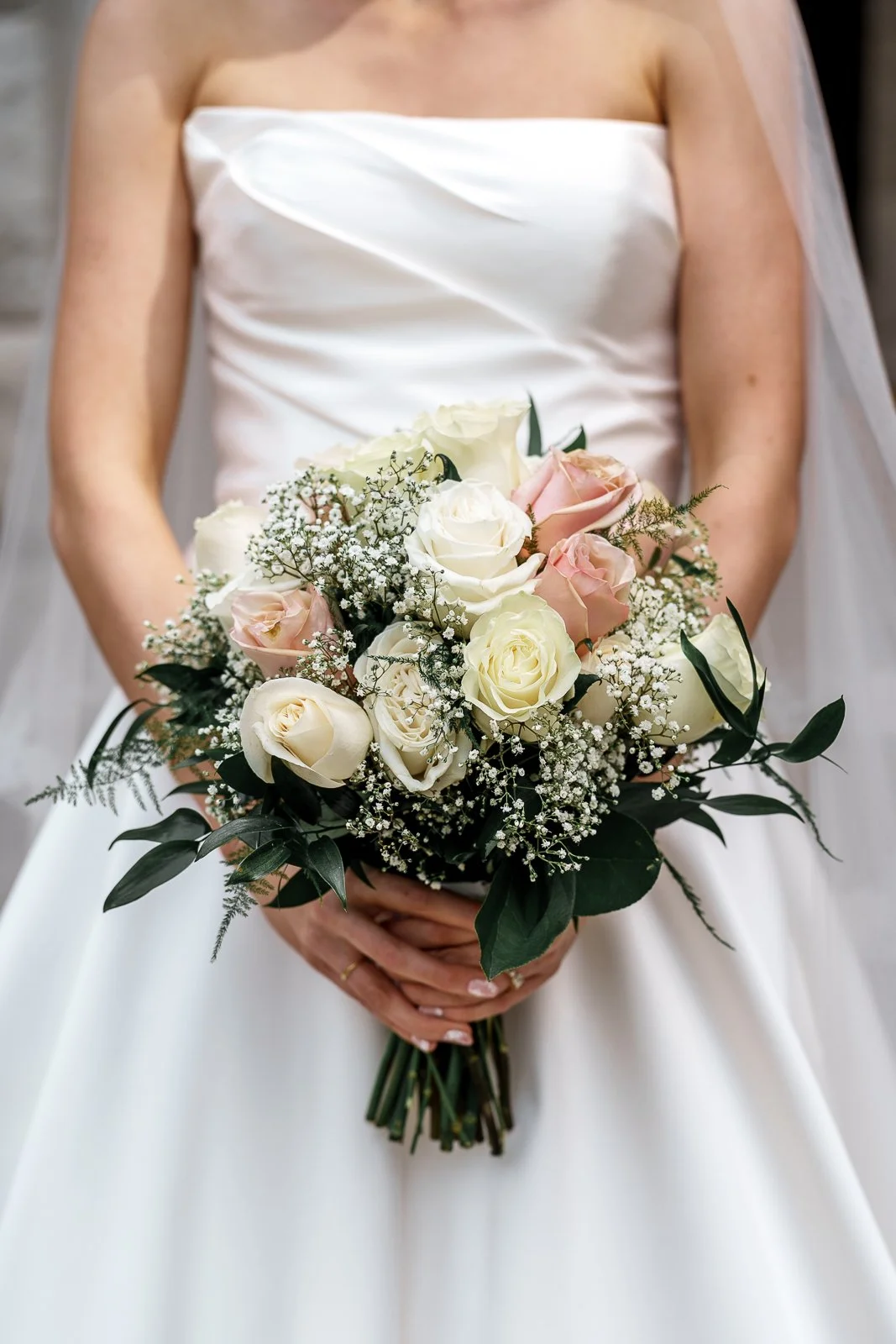 A bride holding a bouquet of white and pink roses with greenery, in front of her white wedding dress.