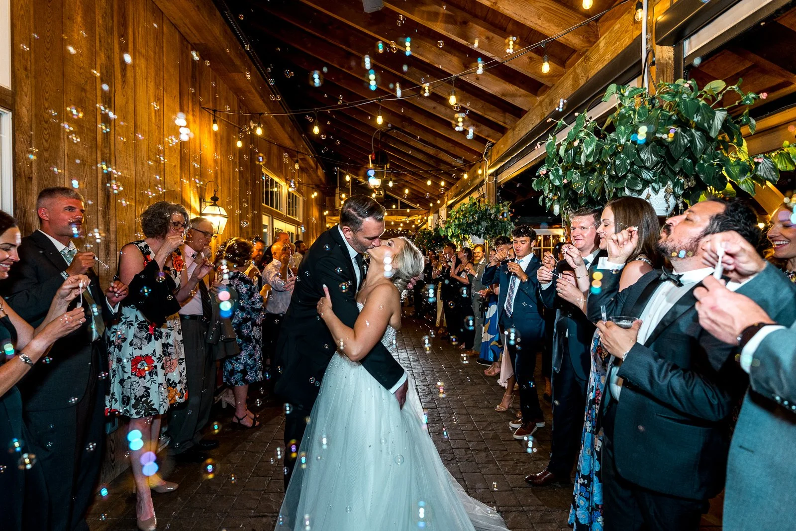 A bride and groom are dancing closely, kissing in the center of a wedding reception with guests on either side celebrating and blowing bubbles under string lights and a wooden ceiling.