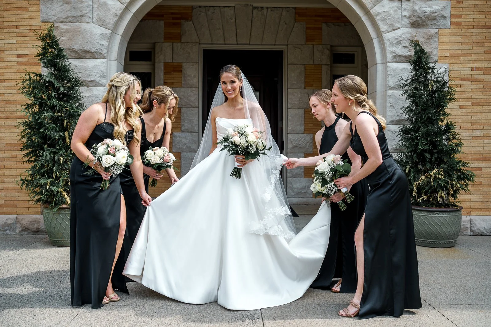 Bride in a white wedding gown holding a bouquet surrounded by bridesmaids in black dresses and holding bouquets, outside in front of a stone and brick building with tall potted plants.
