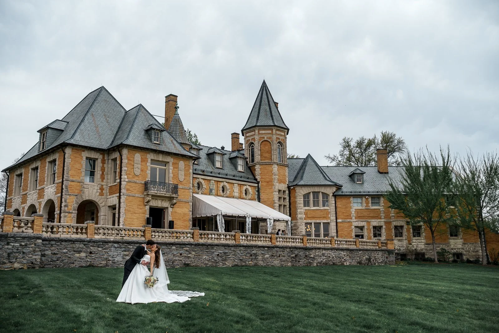A newlywed couple in wedding attire sharing a kiss on a lush green lawn in front of a large, historic mansion with stone and brick architecture, multiple towers, and a covered outdoor area.