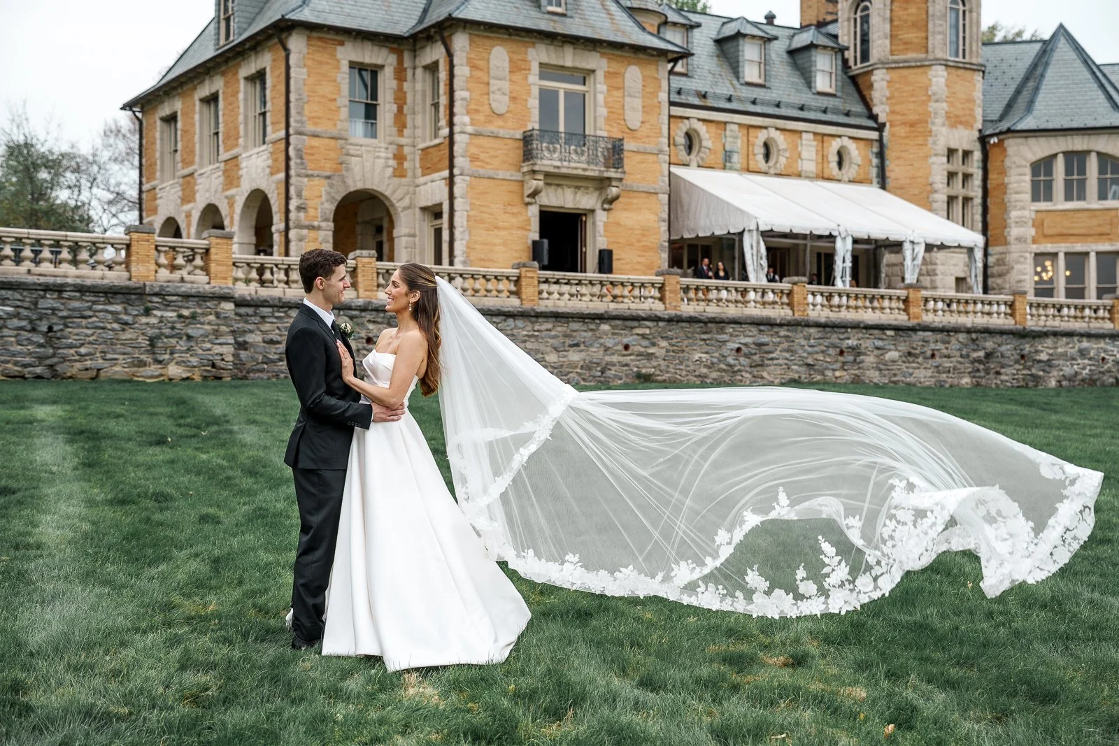 A bride and groom stand on a lush green lawn in front of a large, historic-style house. The bride wears a white wedding gown with a long, flowing veil, and the groom is dressed in a black suit with a white shirt and tie. They face each other, smiling