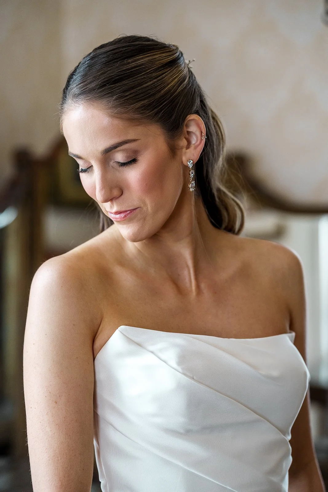 A woman in a white strapless dress with elegant earrings, her brunette hair styled back, with a calm and serene expression, looking downward.