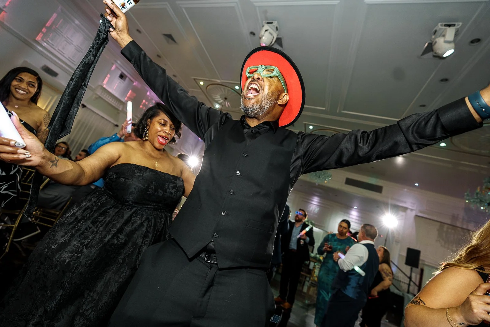 A man wearing a black shirt, a red hat, and colorful glasses dancing energetically at a party, with women in black dresses around him.