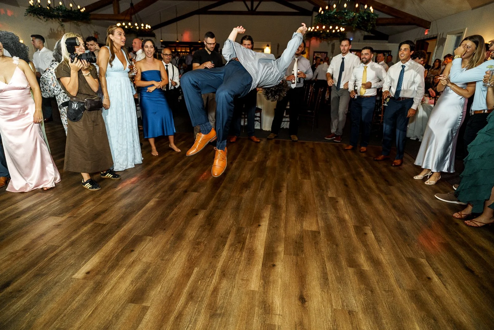 A man in business attire is mid-air doing a backflip on a dance floor surrounded by wedding guests.