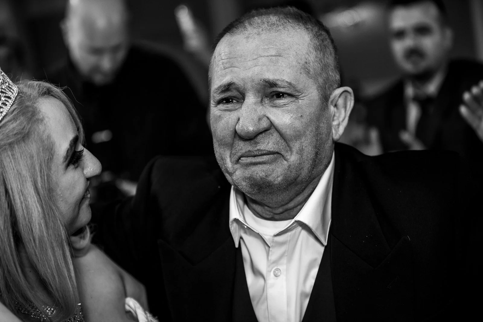 A black and white photo of a man in a suit with a distressed or emotional expression on his face, with a woman and other people blurred in the background.