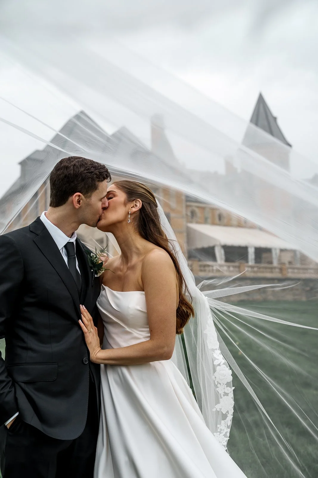 A bride and groom sharing a kiss under a wedding veil outdoors with a historic building in the background.