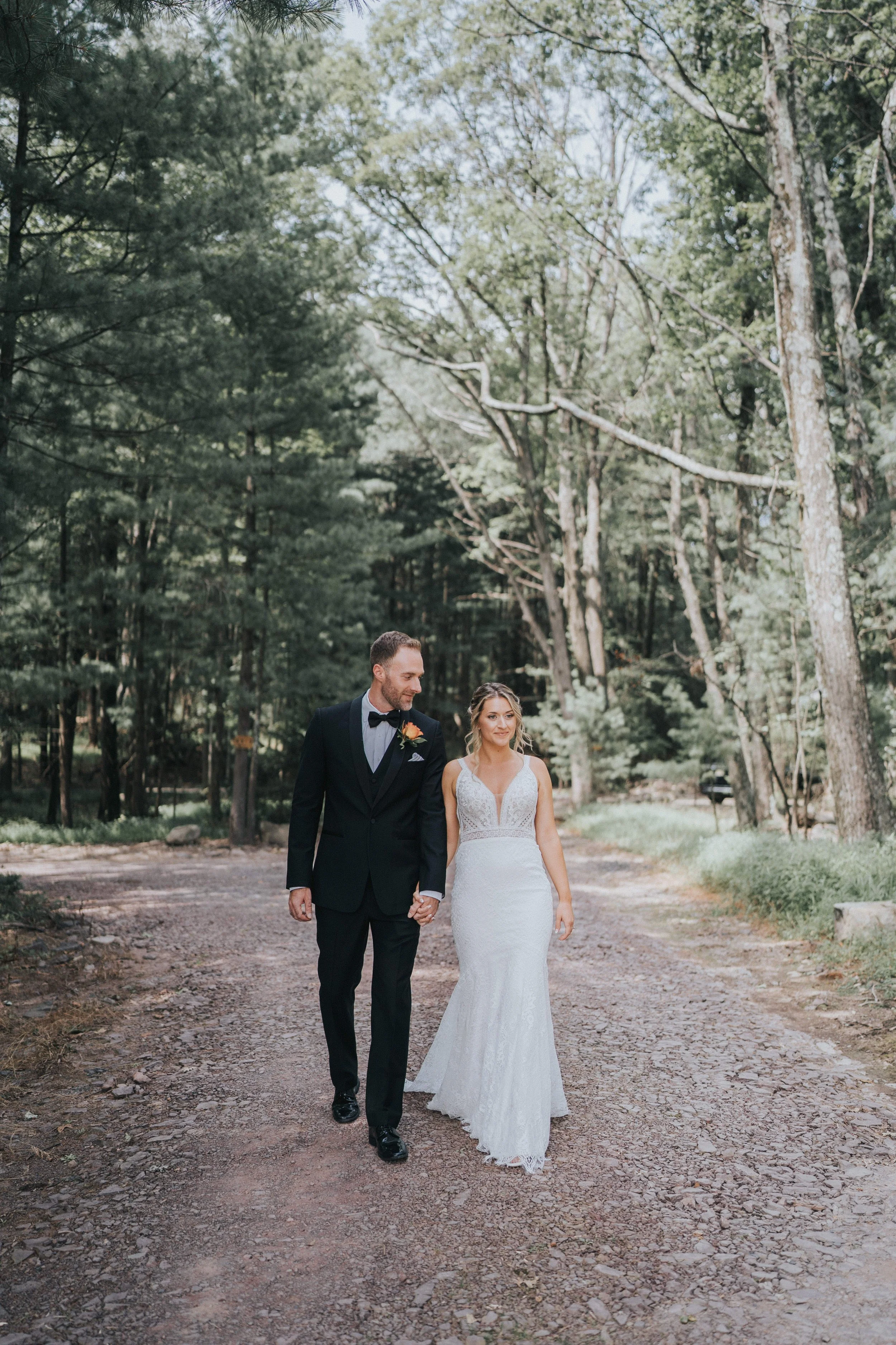 A bride and groom walking hand-in-hand through a wooded outdoor area during their wedding photos, with the bride wearing a white lace gown and the groom in a black tuxedo.