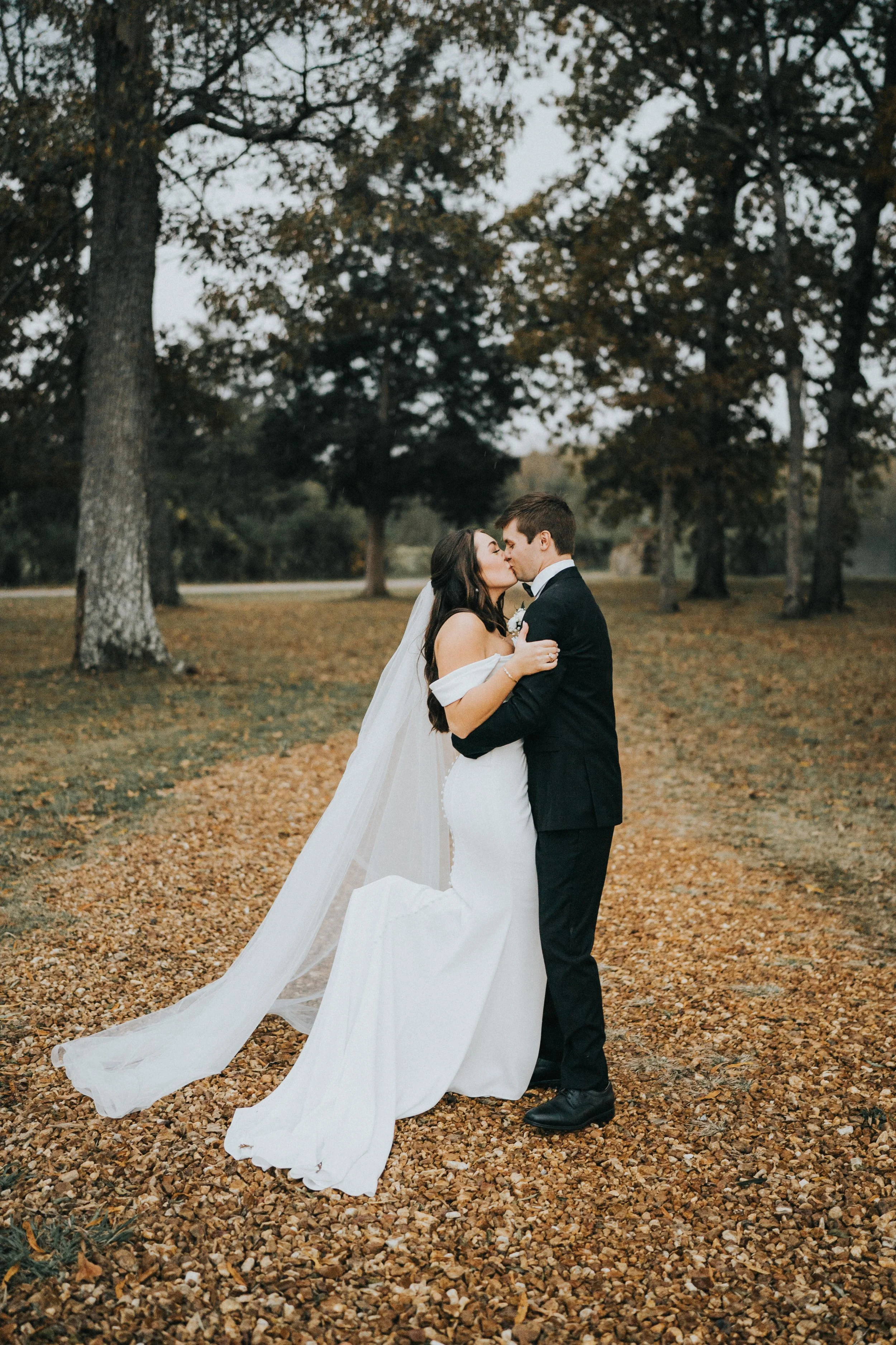 A bride and groom share a kiss outdoors surrounded by trees and fallen autumn leaves.