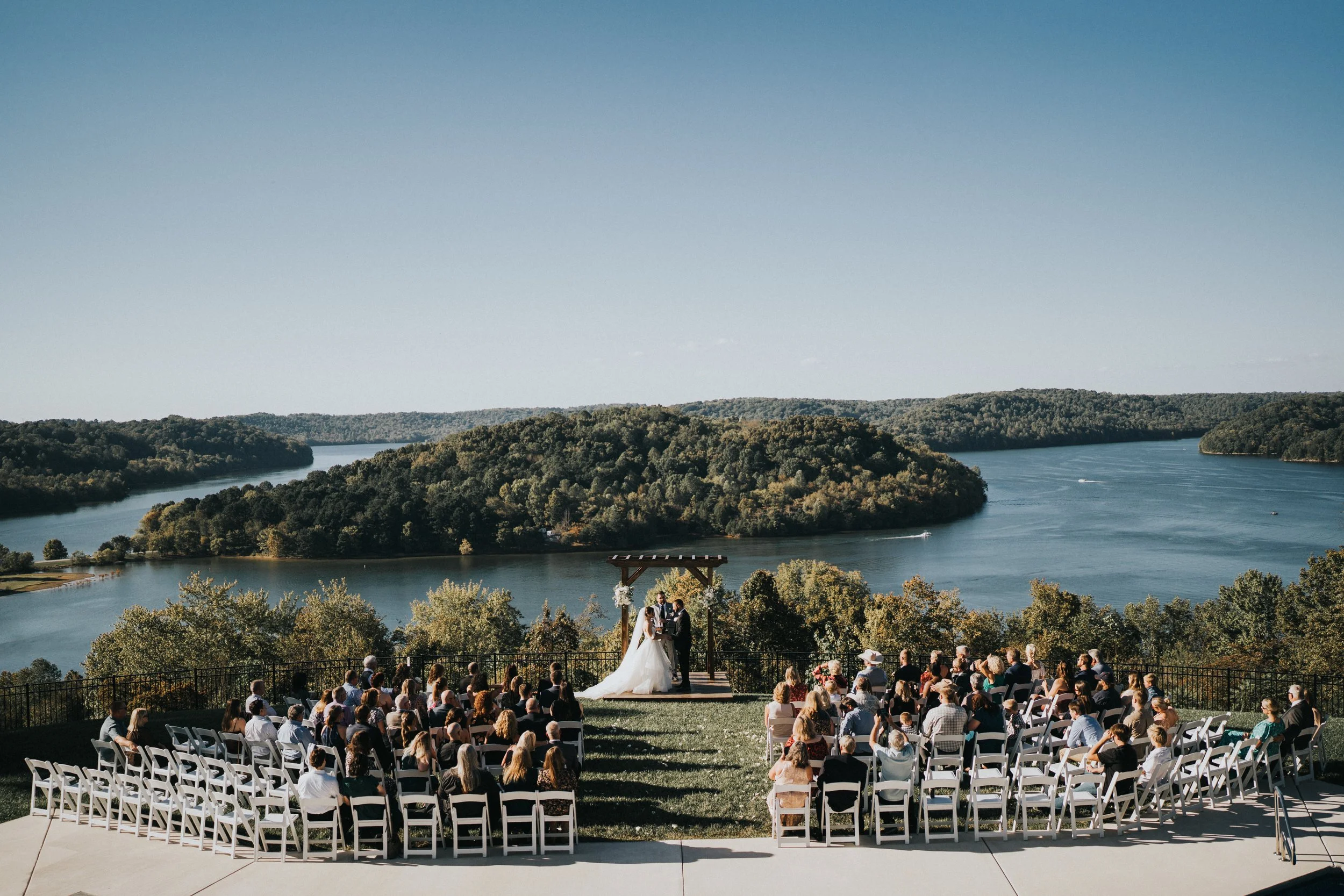 Outdoor wedding ceremony overlooking a lake and forested hills with guests seated in white chairs, couple standing under an arch, during daytime.