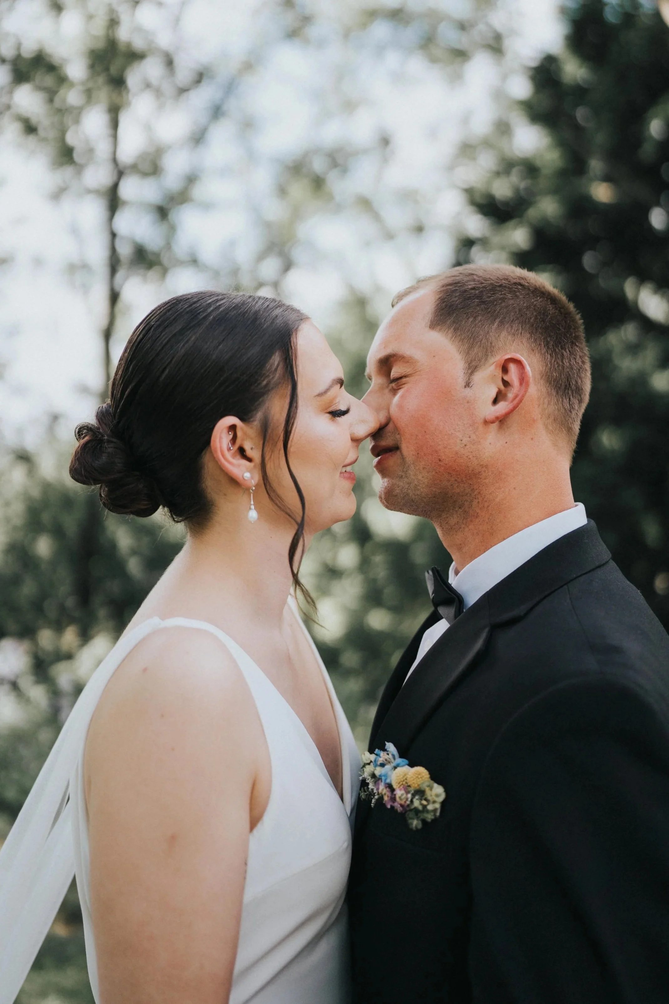 A bride and groom stand face-to-face outdoors with eyes closed, noses touching, wearing wedding attire; the bride in a white dress with pearl earrings and the groom in a black tuxedo with a boutonniere.