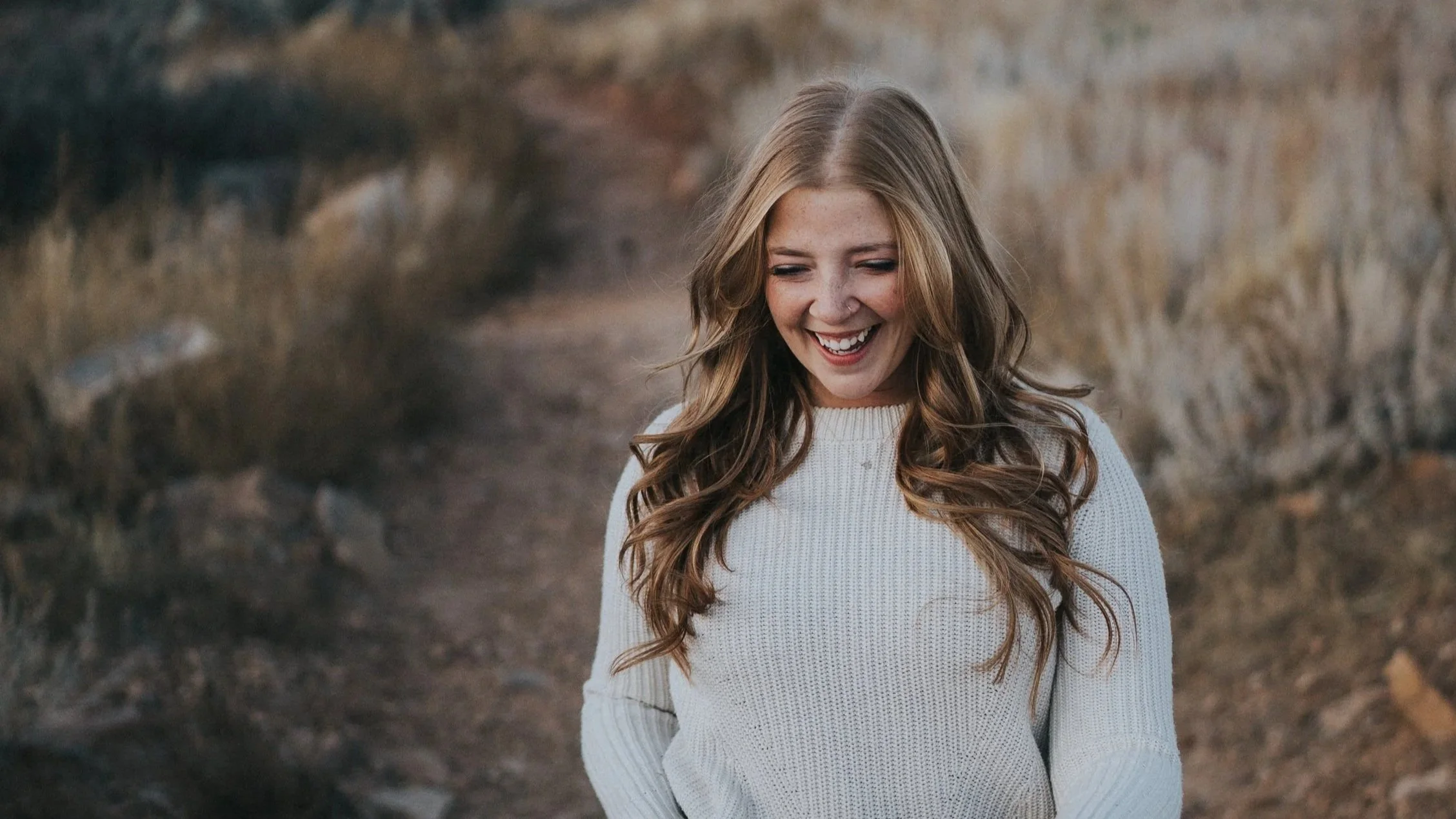 A young woman with long, wavy brown hair smiling and looking down, wearing a white knitted sweater, standing outdoors on a dirt path with dry grass and bushes in the background.