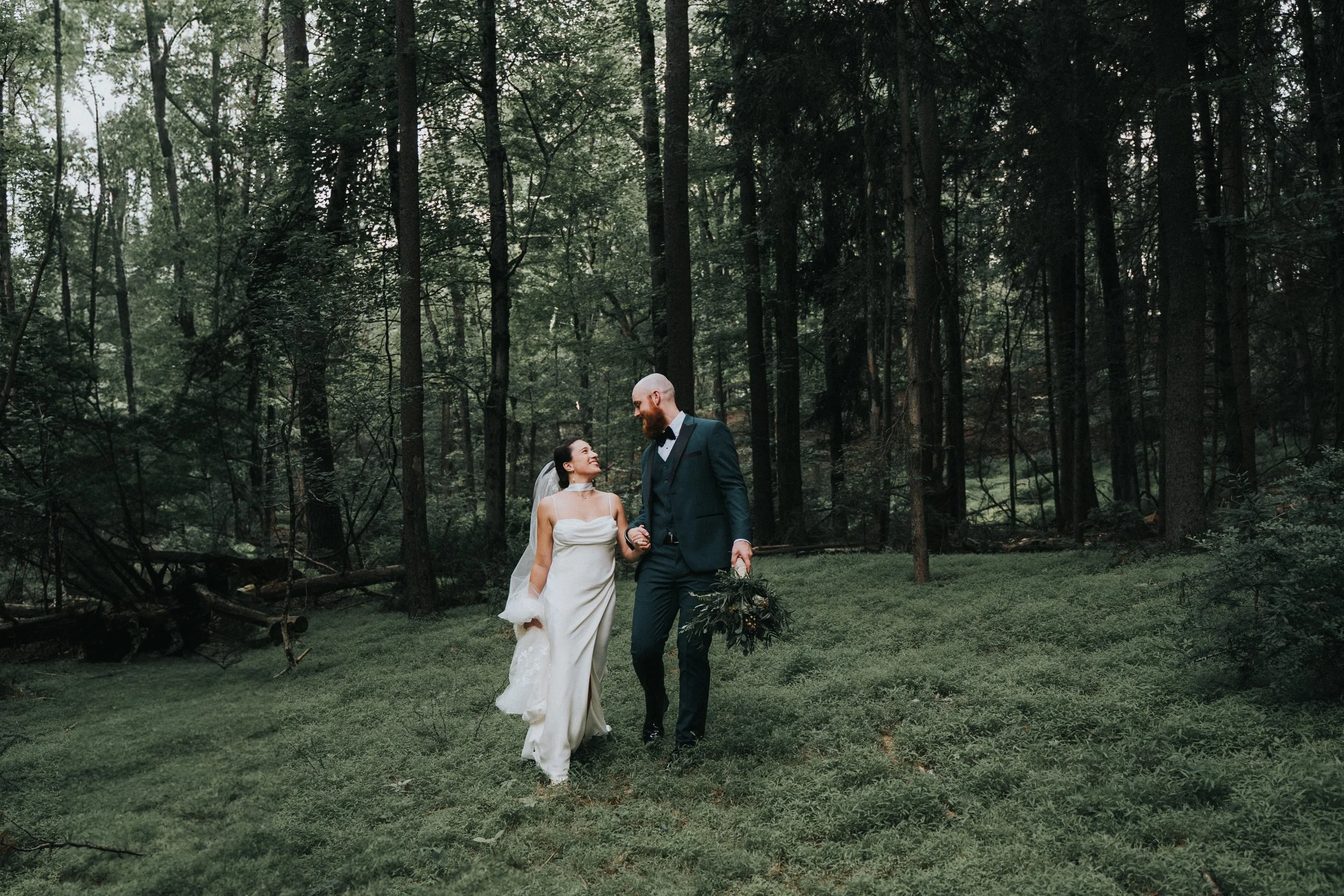 A bride and groom walking hand in hand through a forested area during their wedding, with the bride in a white dress and the groom in a dark suit holding a bouquet.