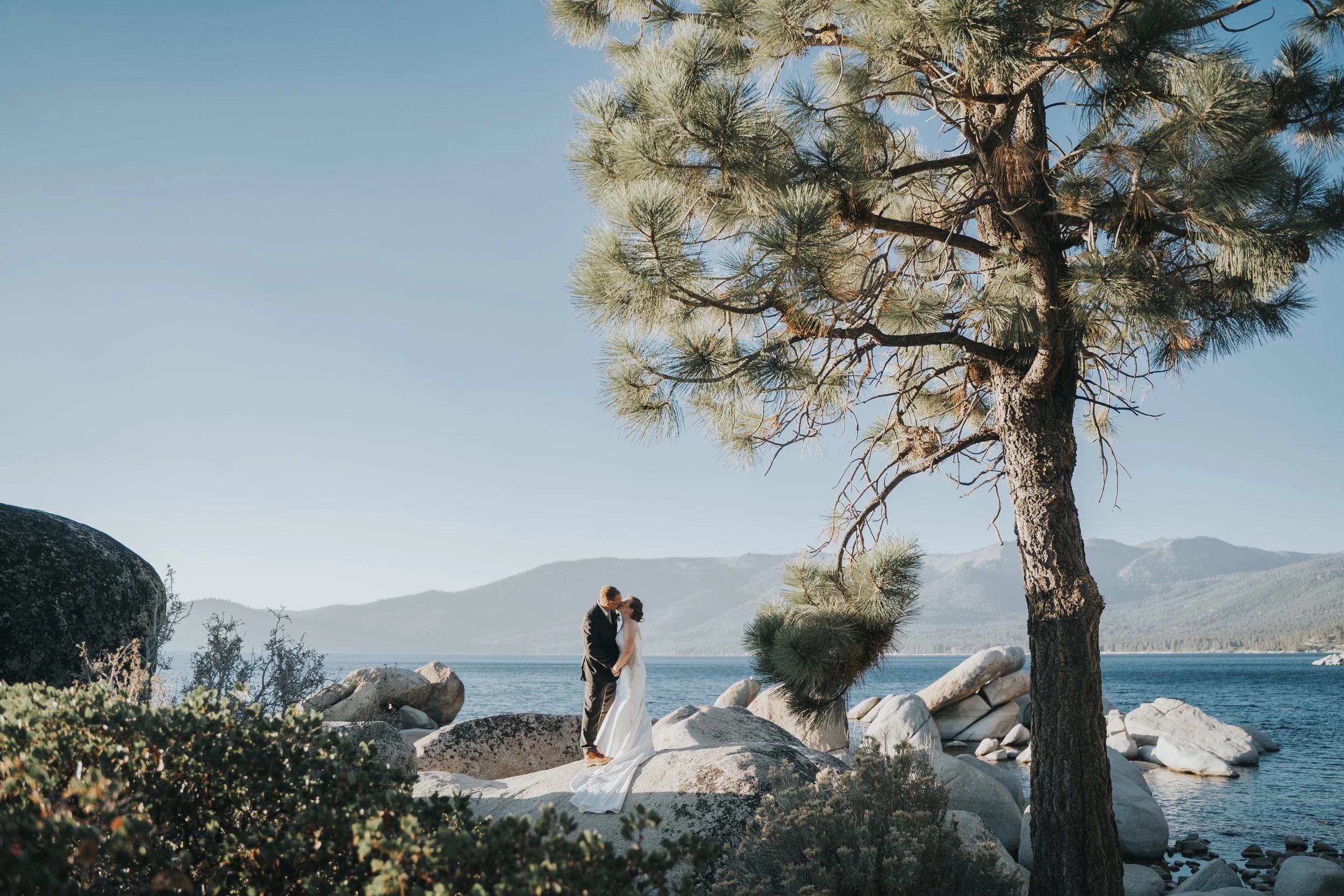 A bride and groom standing on rocks by a lake, kissing, with a pine tree and distant mountains in the background.