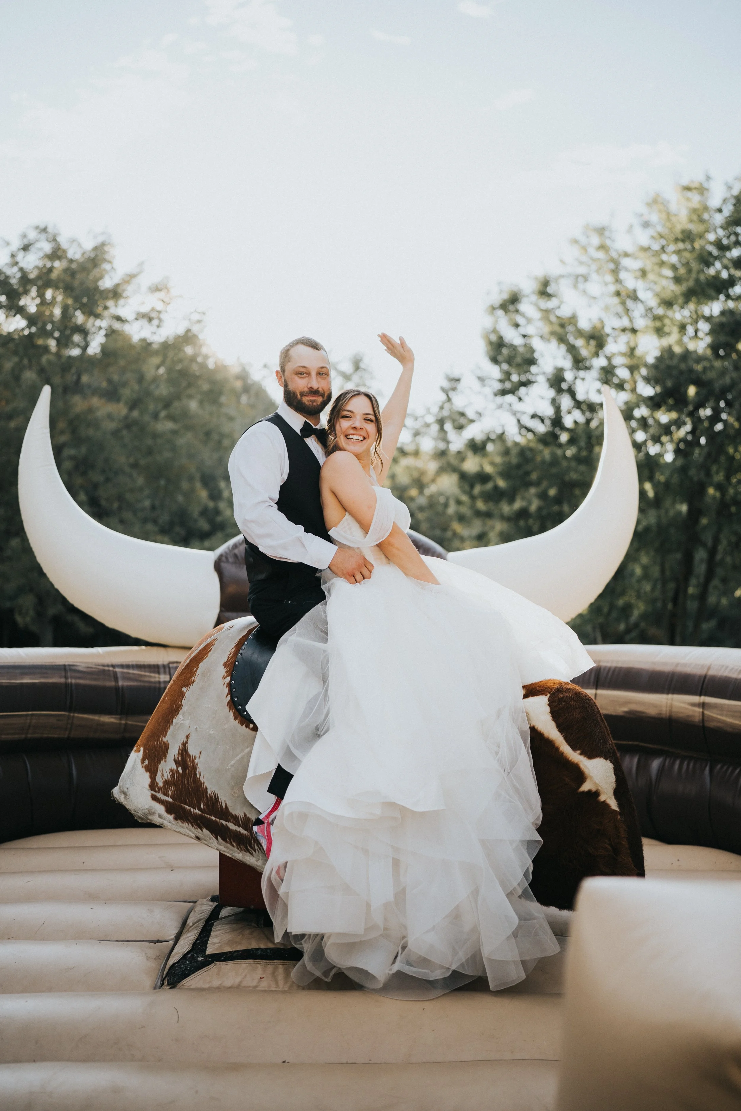 A bride and groom riding a mechanical bull shaped like a cow, with large crescent moon decorations in the background during a wedding celebration outdoors.