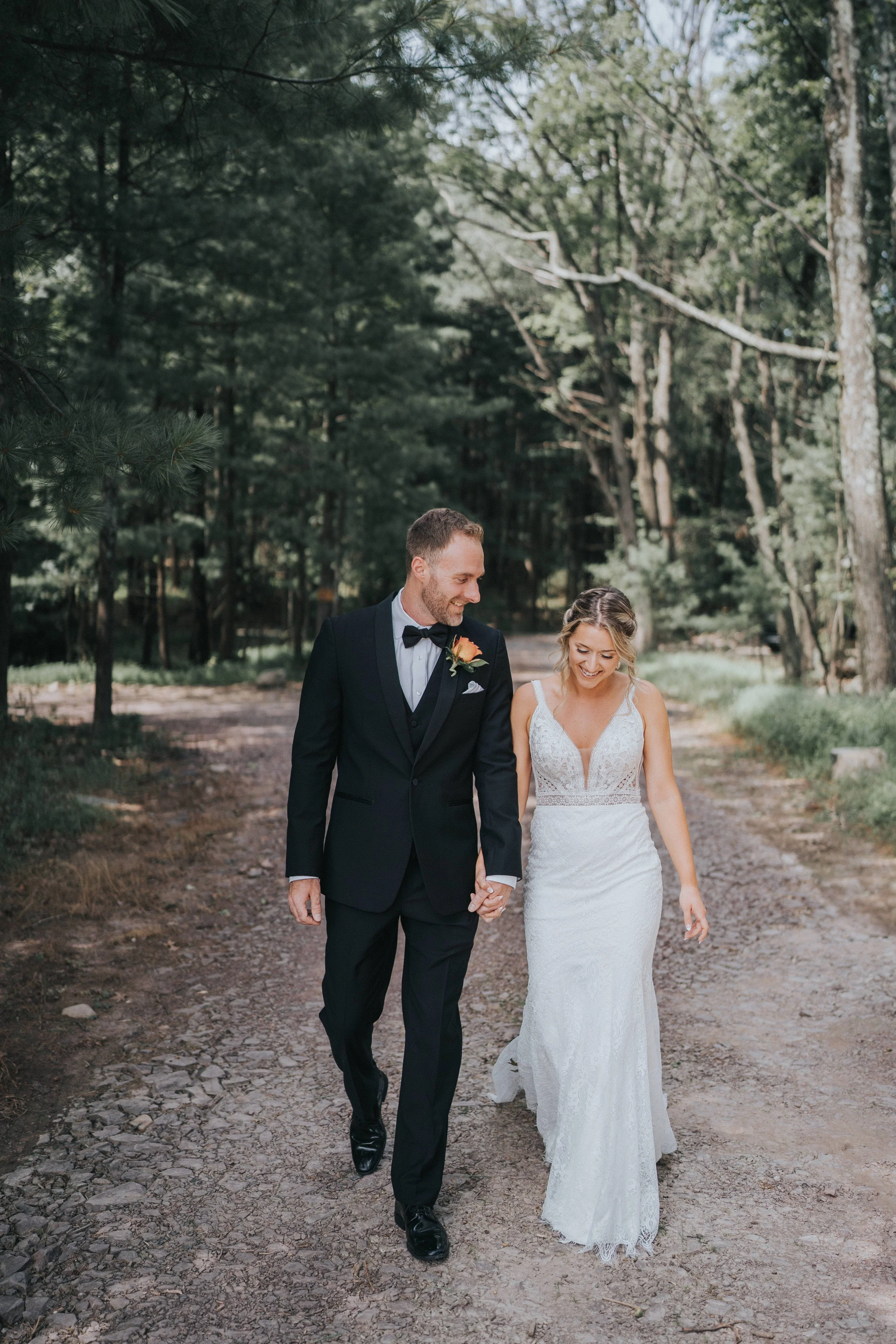 A newlywed couple walking hand-in-hand through a wooded area on their wedding day, dressed in formal wedding attire.