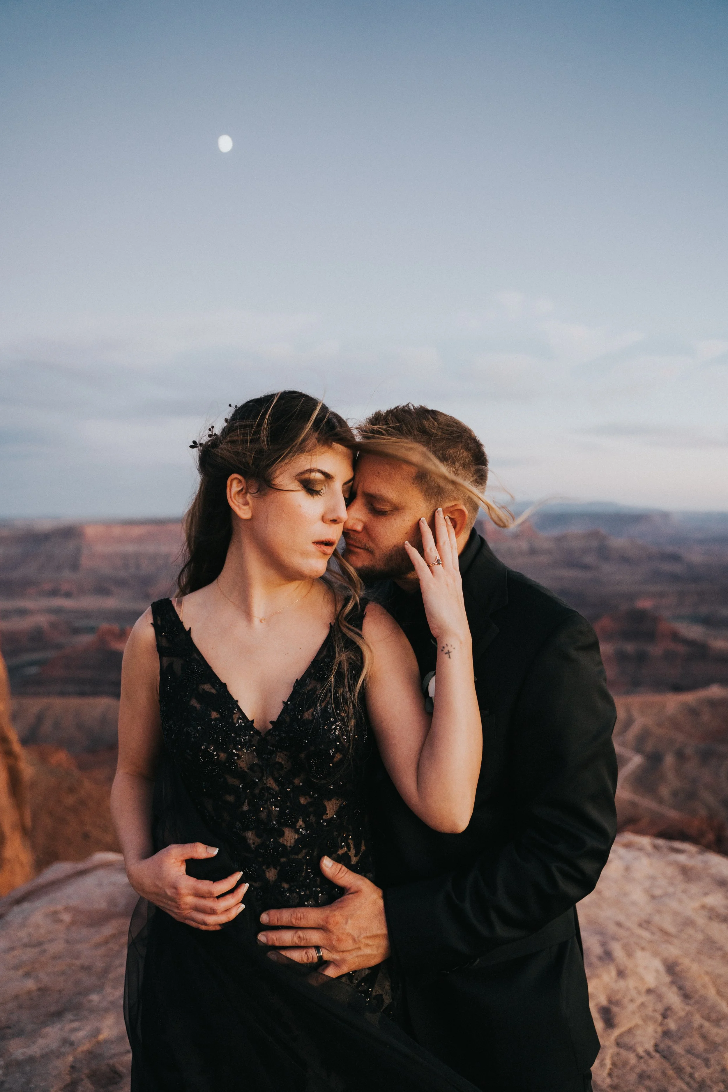 A couple embracing on a rocky ledge at sunset with a view of canyon formations in the background, moon visible in the sky.