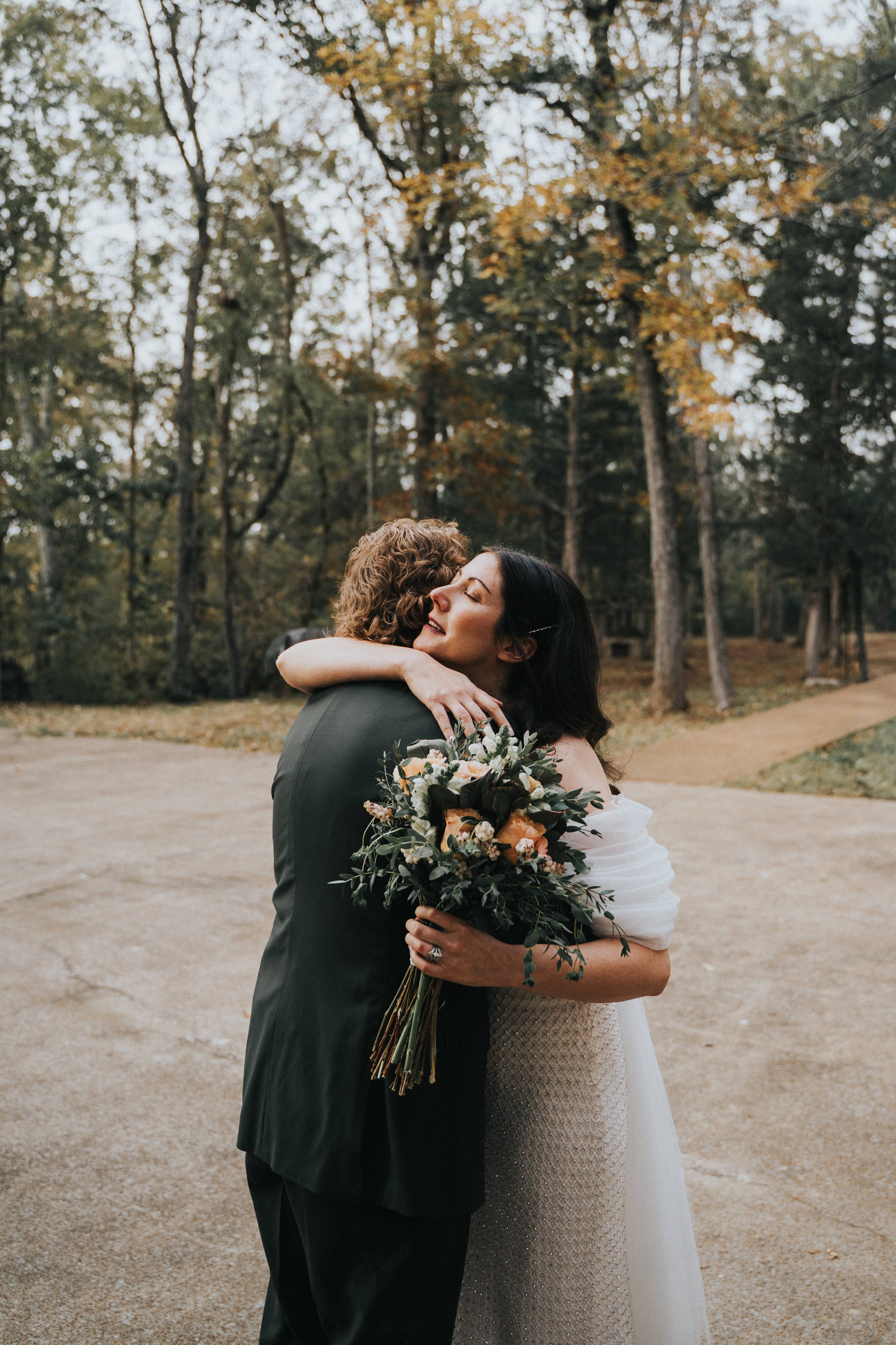 A bride and groom hugging outdoors in a wooded area, with the bride holding a bouquet of flowers.