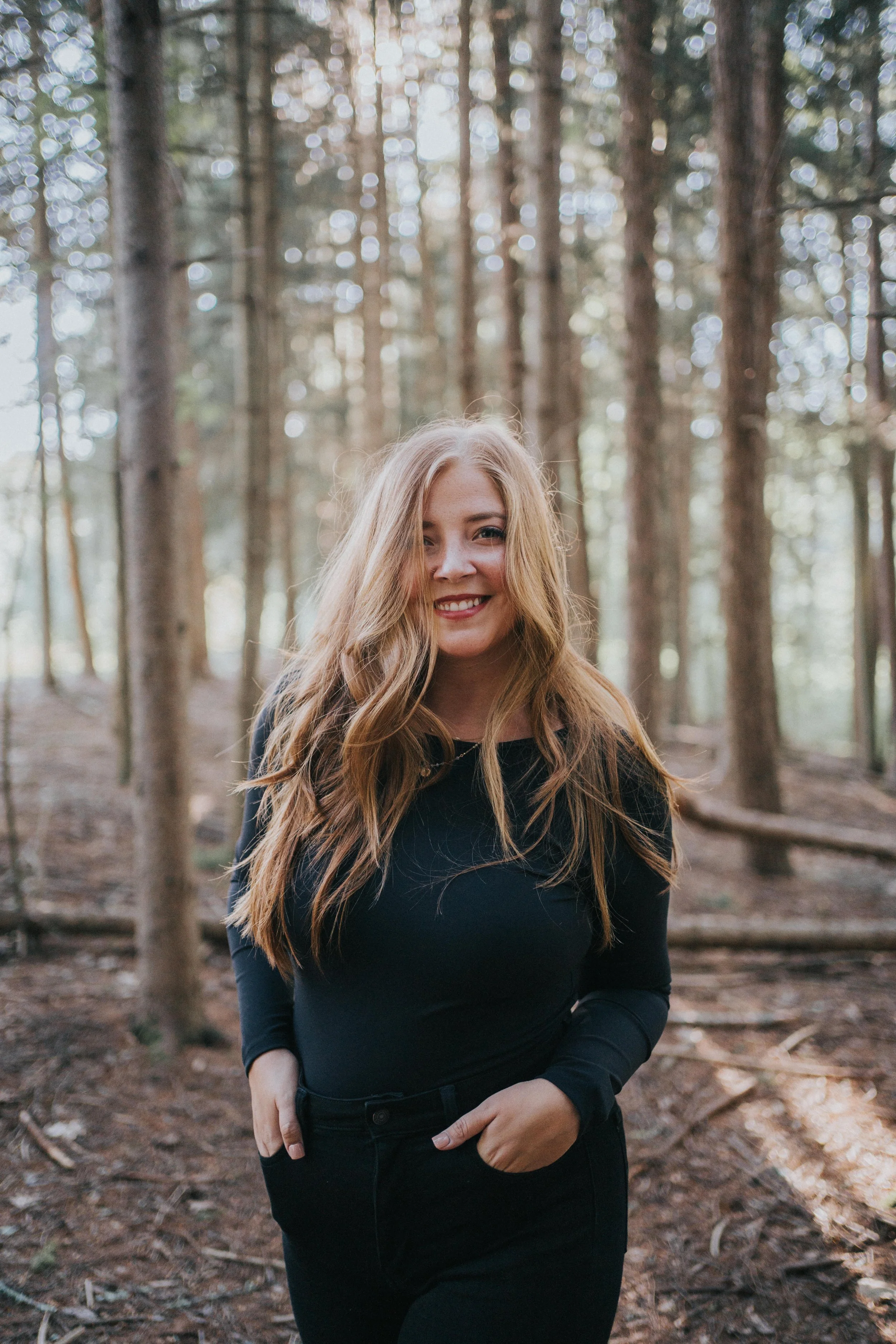 A woman with long wavy red hair smiling and standing in a forest with tall trees and dappled sunlight.