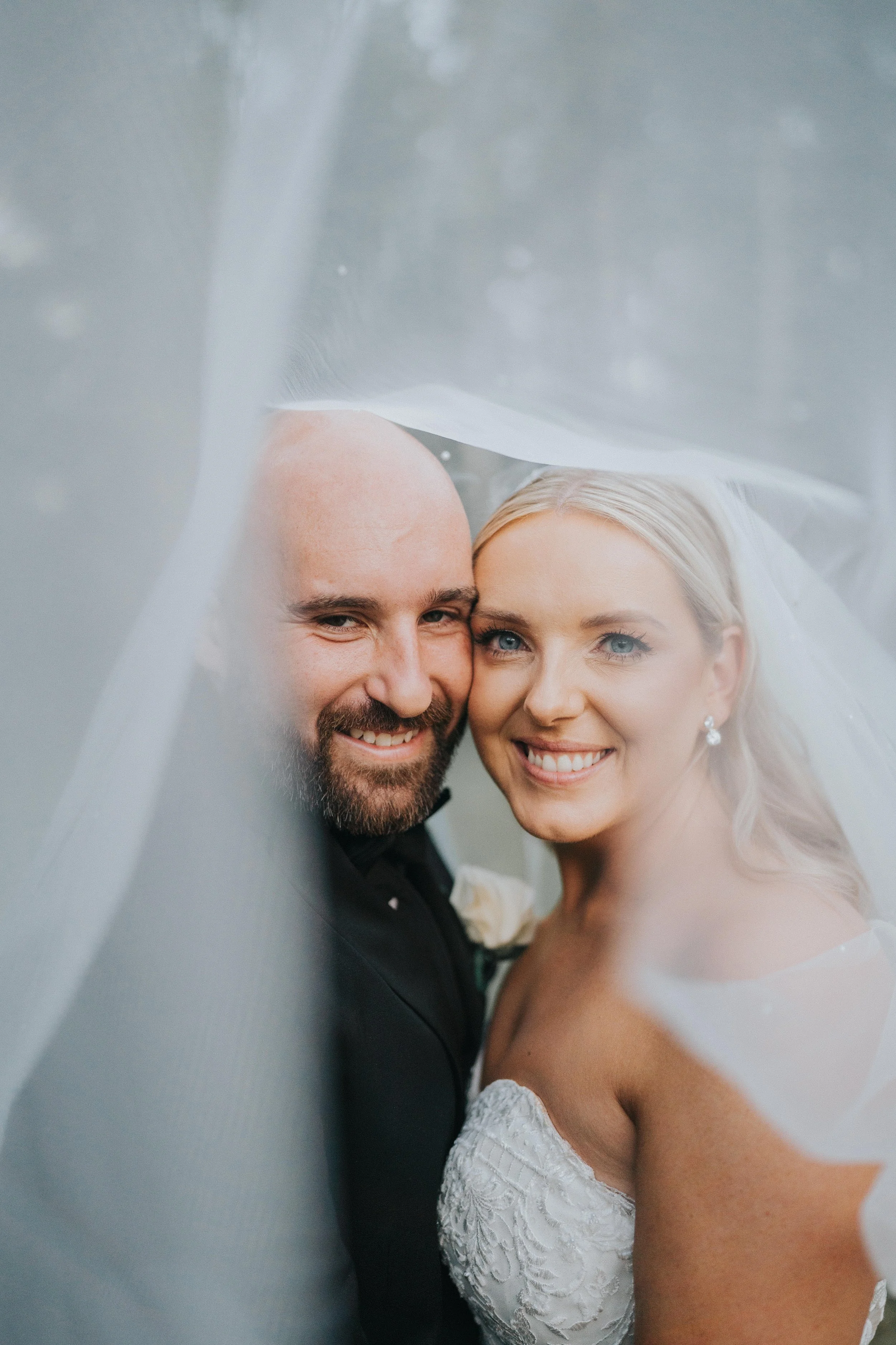 Close-up of a smiling bride and groom on their wedding day, partially covered by a wedding veil.