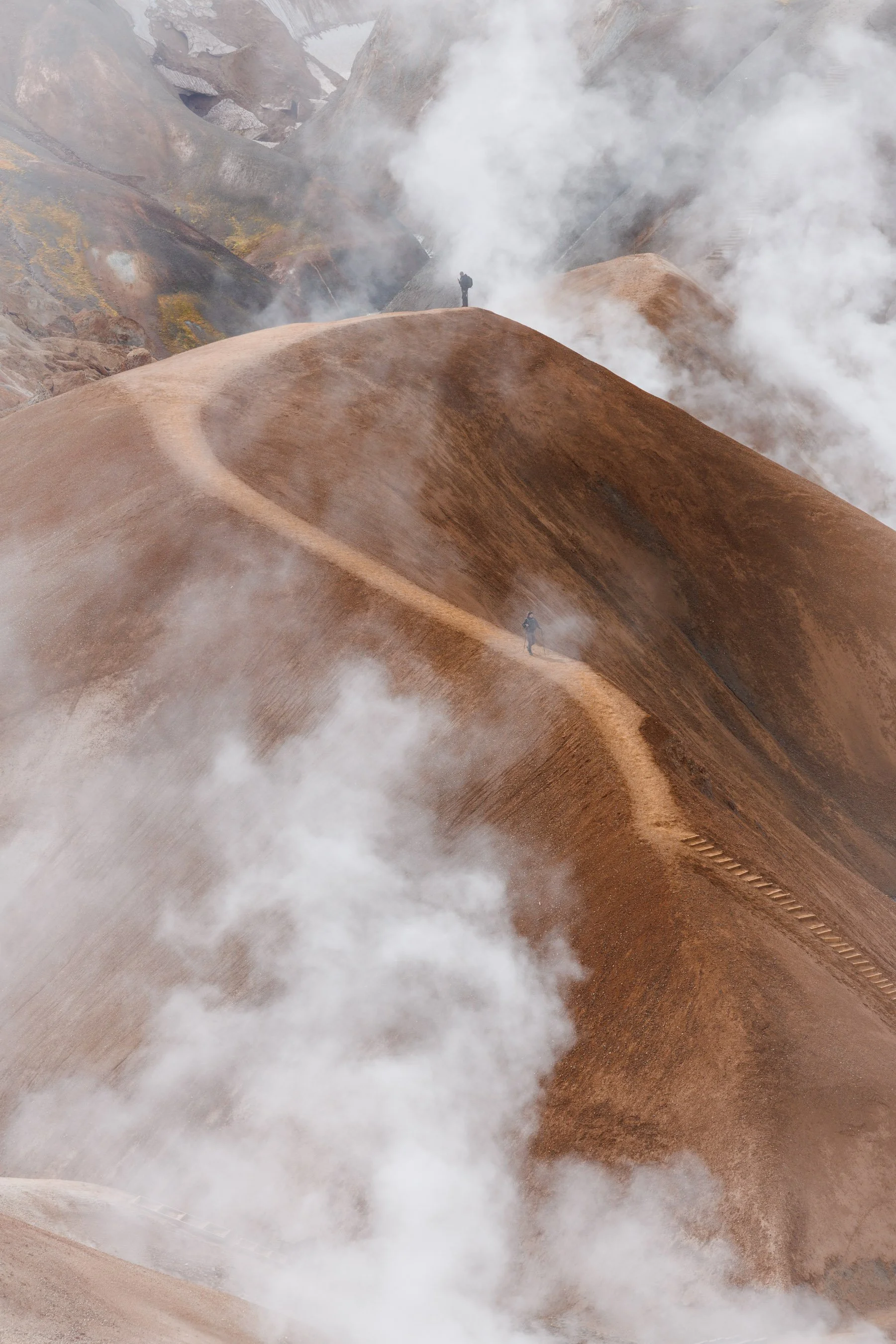 Two hikers walk along a narrow ridge trail through a steaming geothermal landscape, kerlingarfjöll,  surrounded by thick white mist and rust-colored volcanic hills.