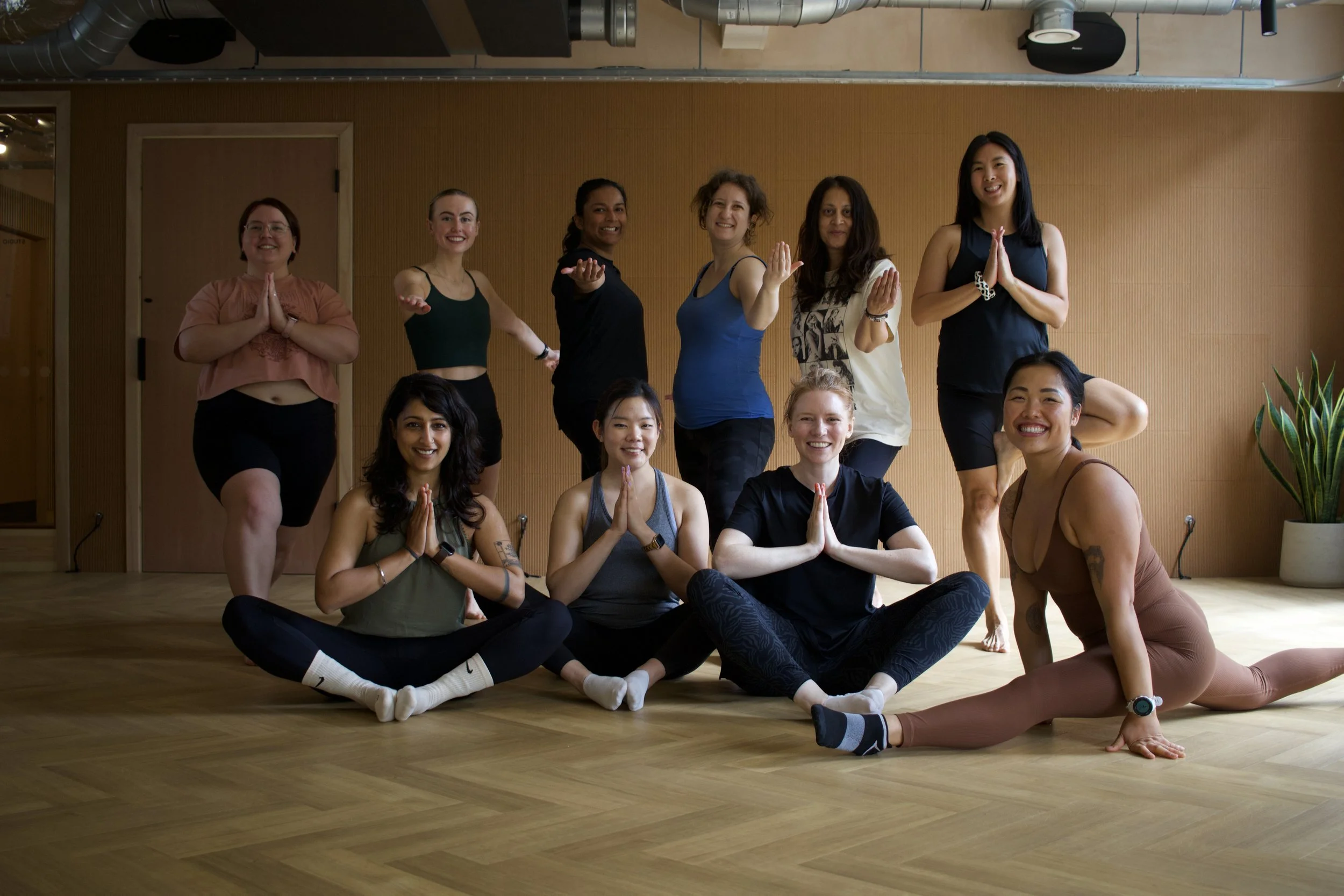 Group of ten women practicing yoga in a studio, some sitting cross-legged on the floor with hands in prayer position, and others standing with hands in a prayer pose, smiling and relaxed.