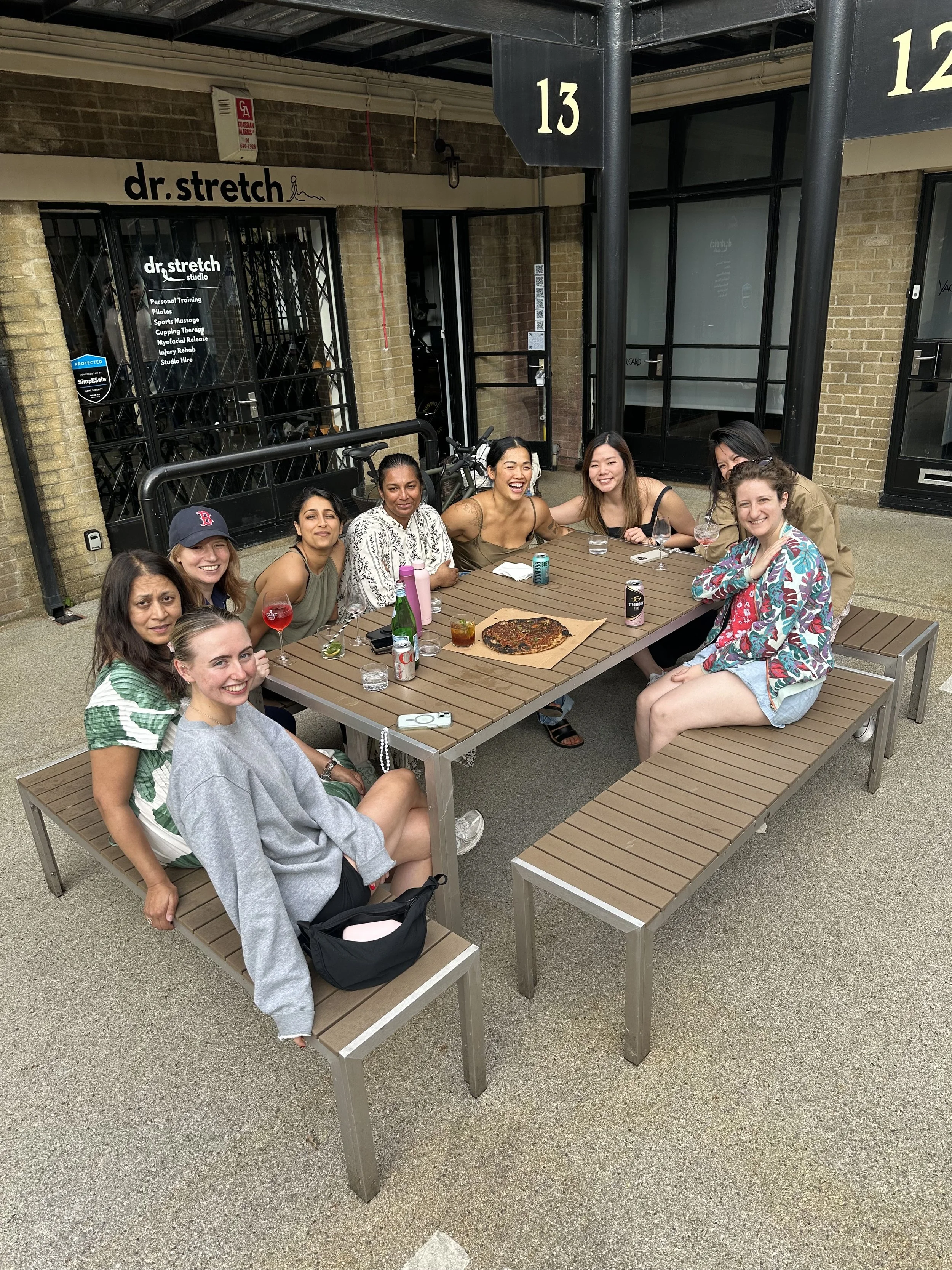 Group of eight women sitting around a wooden outdoor table, smiling and enjoying drinks and pizza, in front of a building with signs that read 'dr.stretch'.