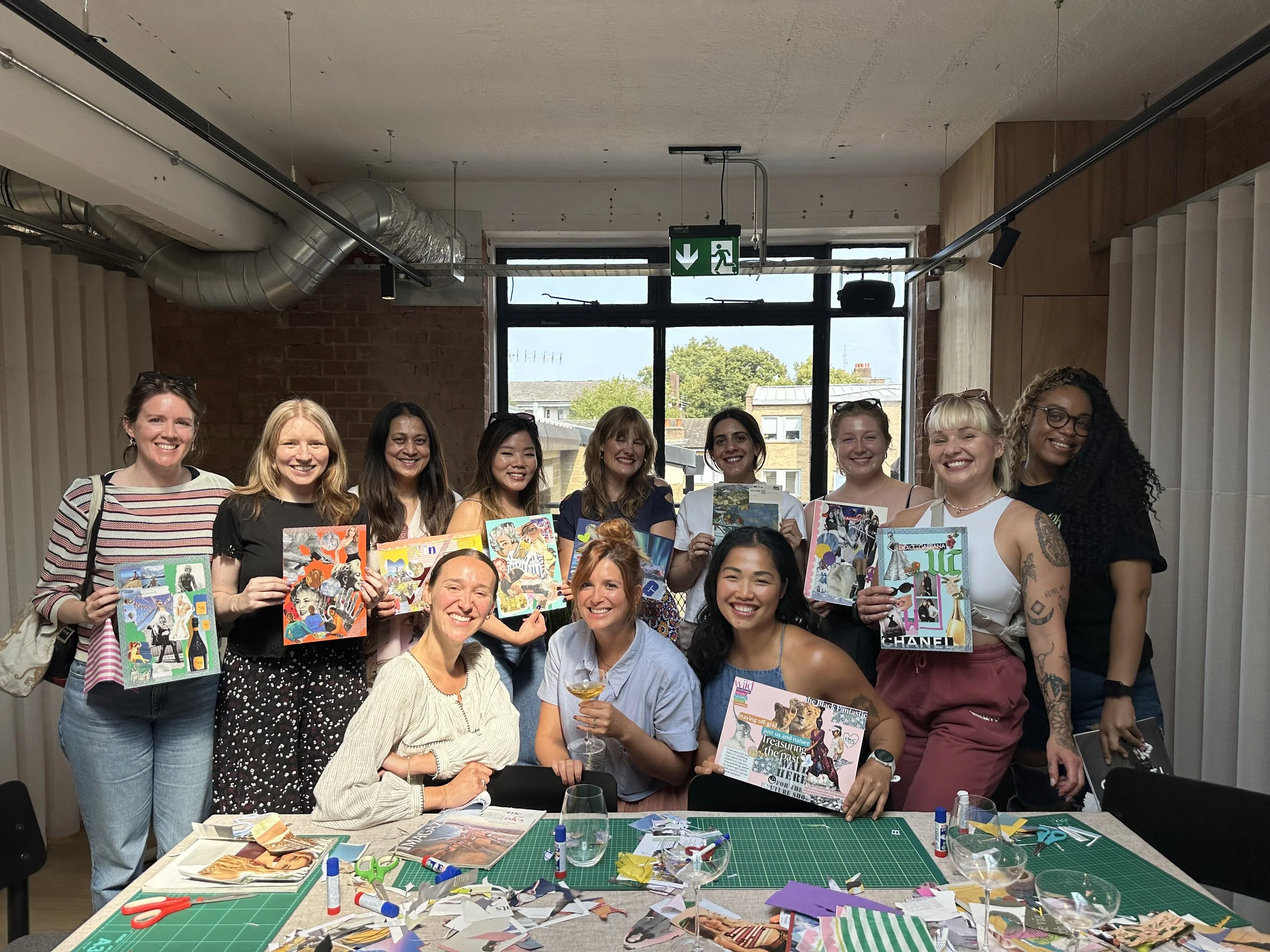 Group of women holding colorful artwork at an art workshop, with craft supplies on the table in front of them.