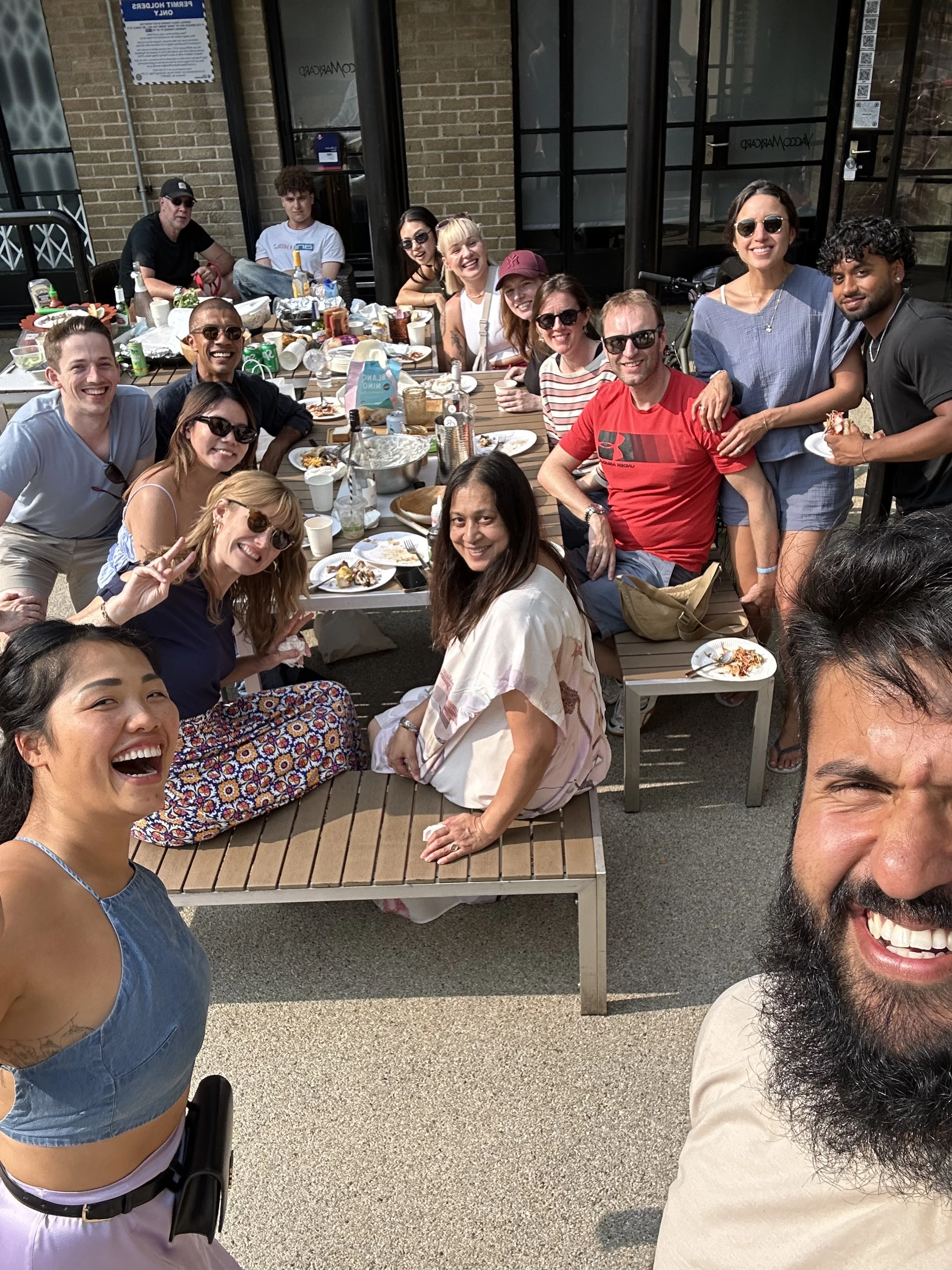 A group of people enjoying a meal together outdoors, sitting at a table filled with food and drinks, smiling and taking a selfie.