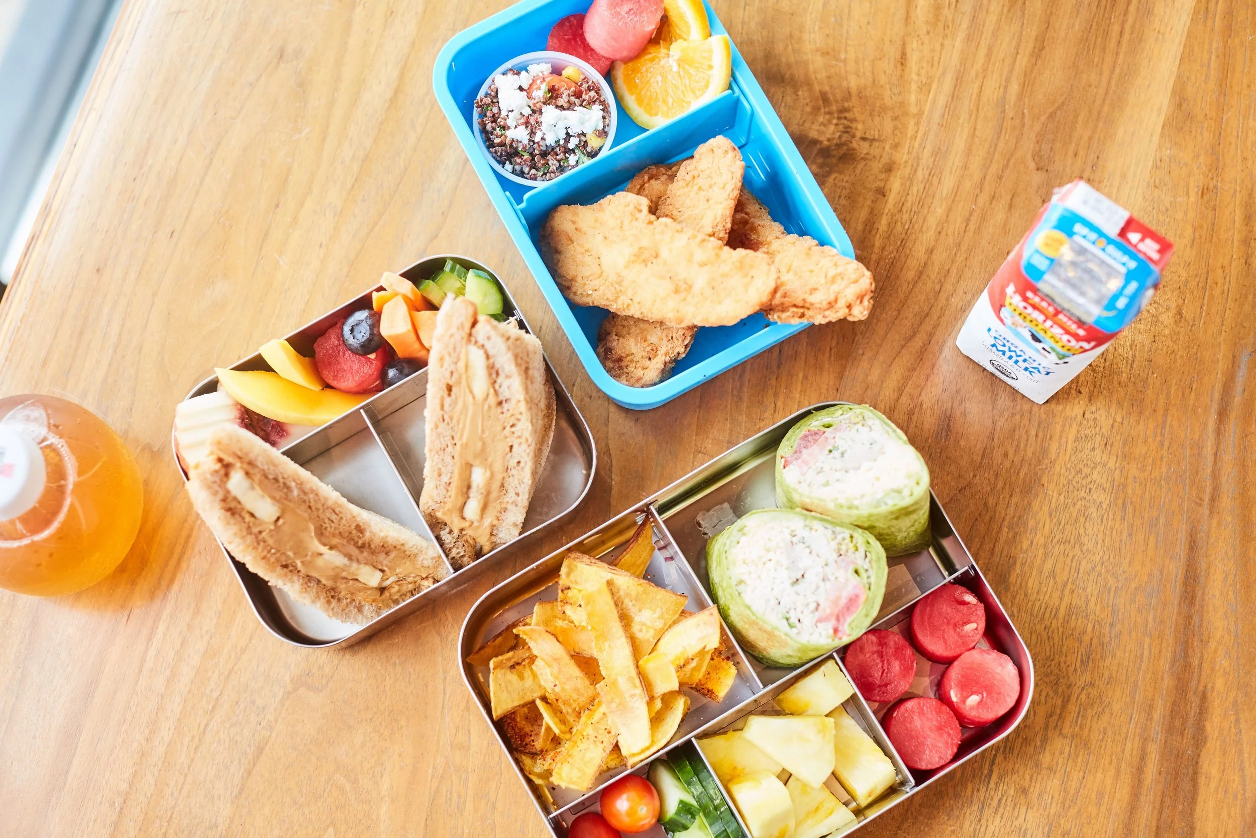 Baked fish fillets with crispy breading, potato chips, fried plantains, and a side of creamy dipping sauce in a metal container, served on red and white checkered paper in a metal basket.