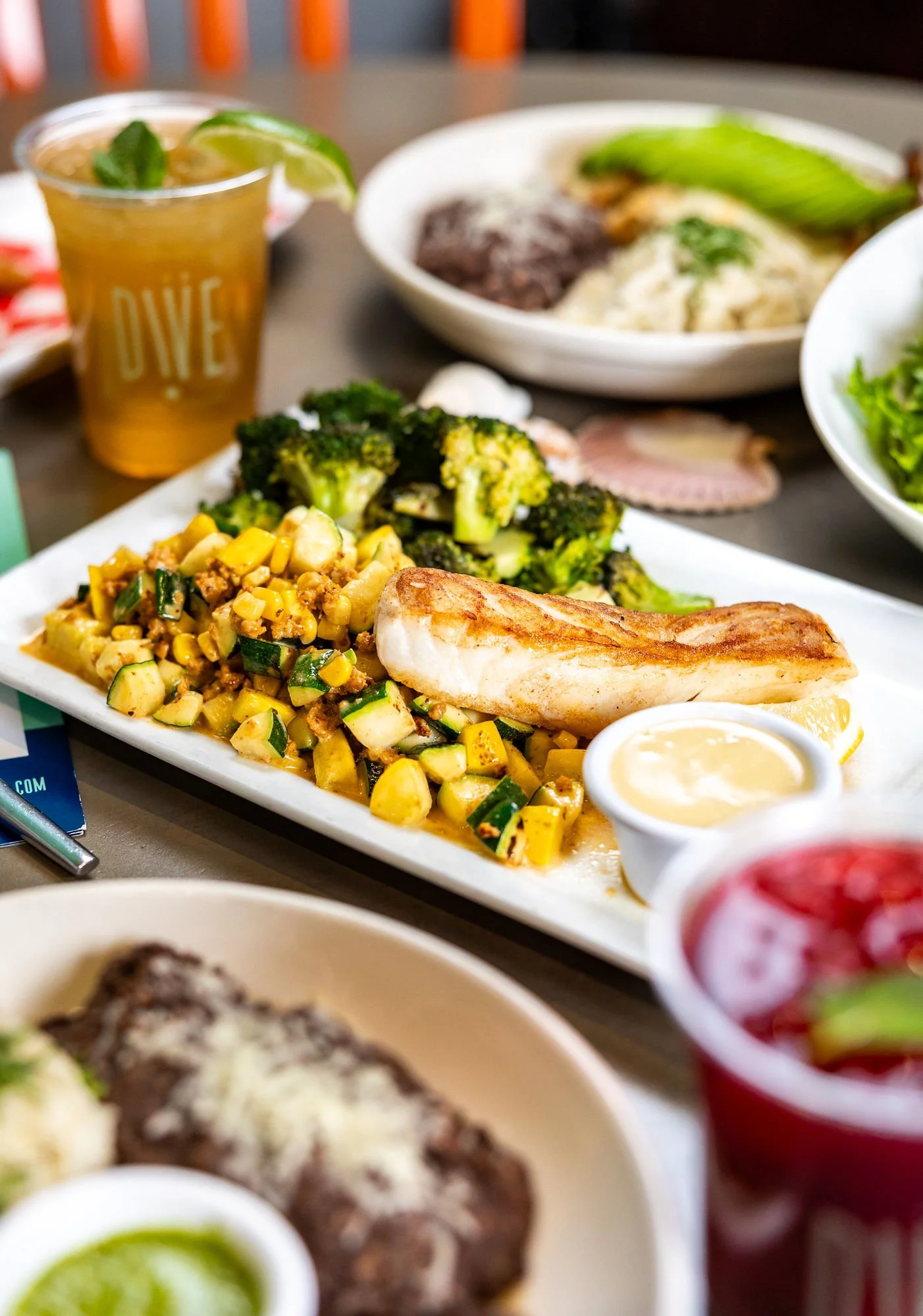 A dinner plate with grilled fish, roasted asparagus, mashed potatoes, lemon slices, and a side of creamy sauce, alongside a glass of white wine on a wooden table.