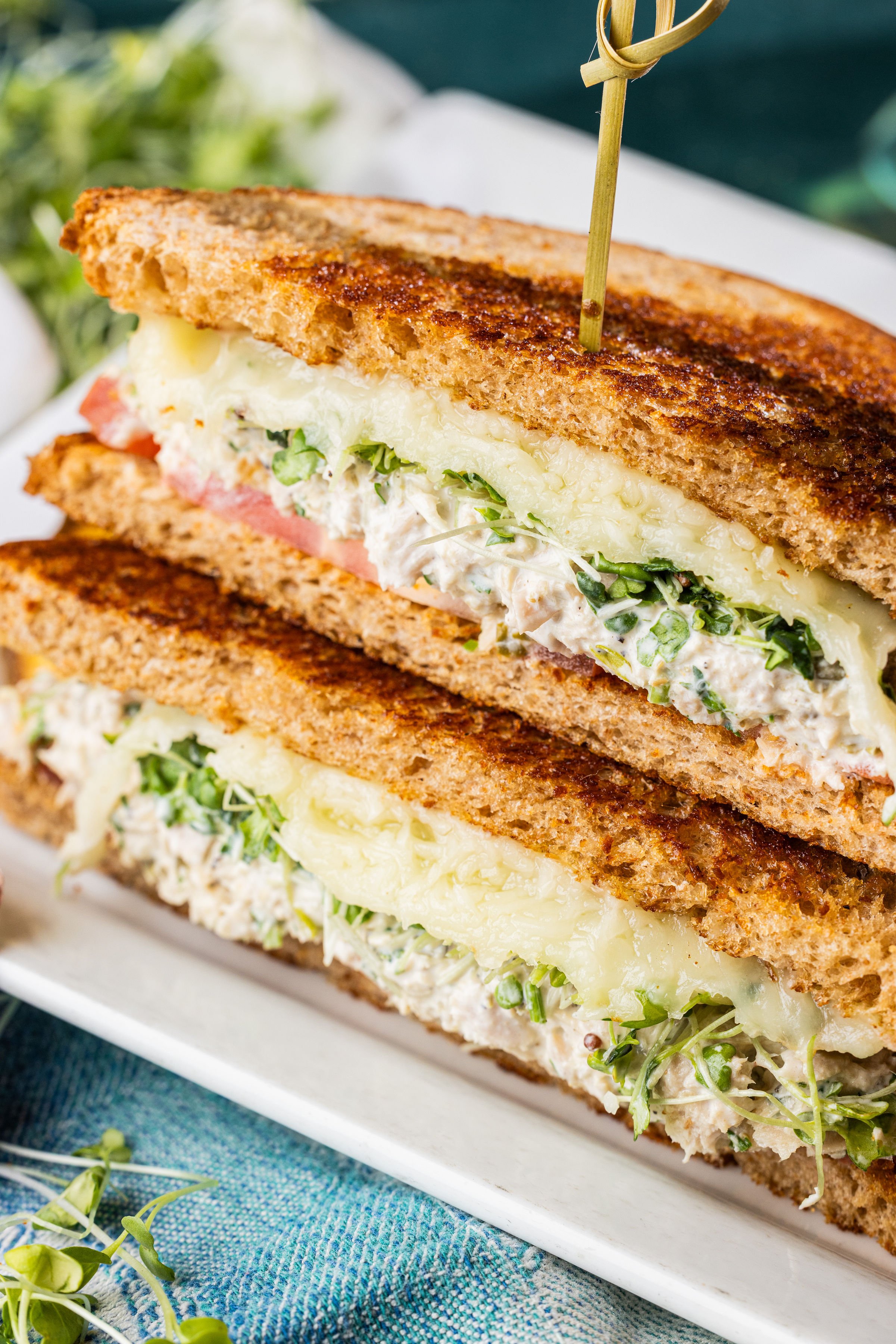 Triple-decker sandwich with lettuce, tomato, and sauce on a plate, served with potato chips and dipping sauce in a small white bowl, on a wooden table in a restaurant.