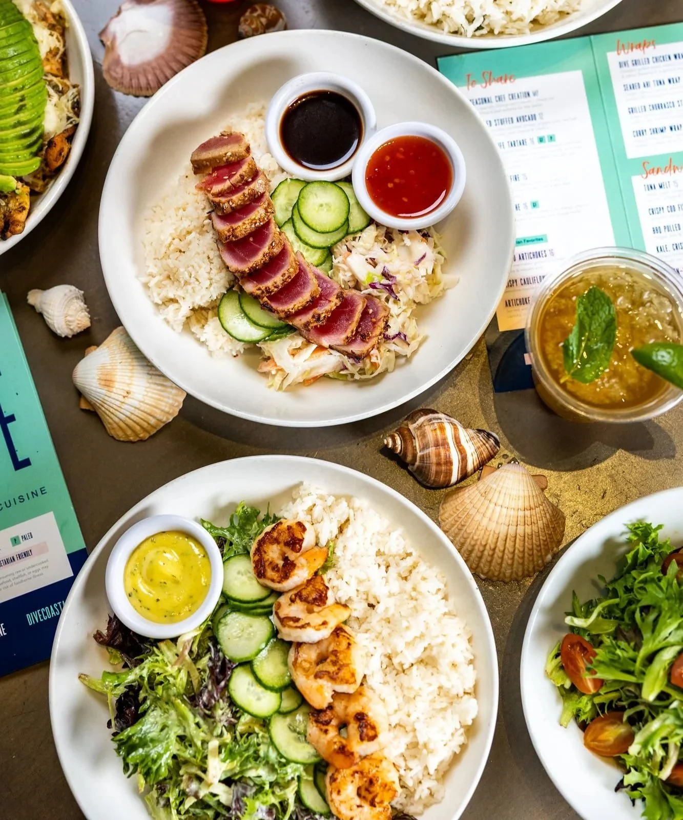 A colorful spread of food including a salad with avocado and blueberries, a taco, a burger, a glass of red drink with fruit, a small bowl of fruit, and a plate of grilled meat, all on a blue-striped tablecloth with sunflower decorations.