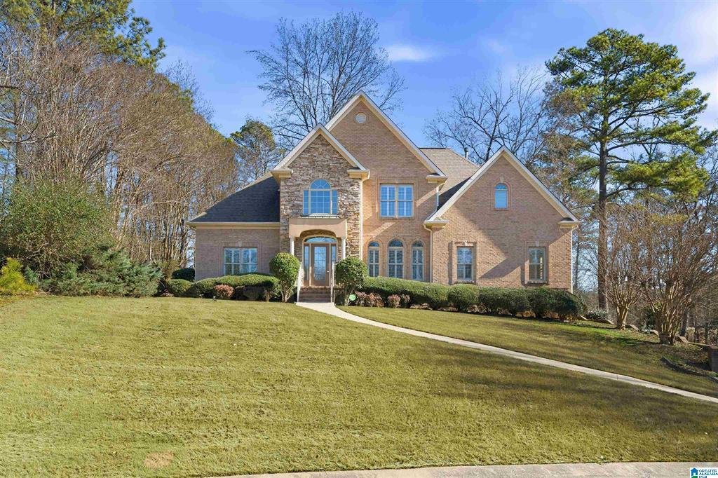A large brick house with a steep roof, situated on a sloped lawn with some trees in the background, under a partly cloudy sky.