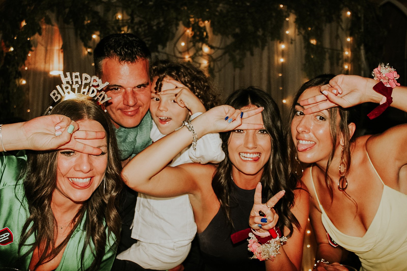Grupo de personas celebrando en una fiesta con decoraciones de luces y fondo oscuro, haciendo gestos divertidos y saludando.