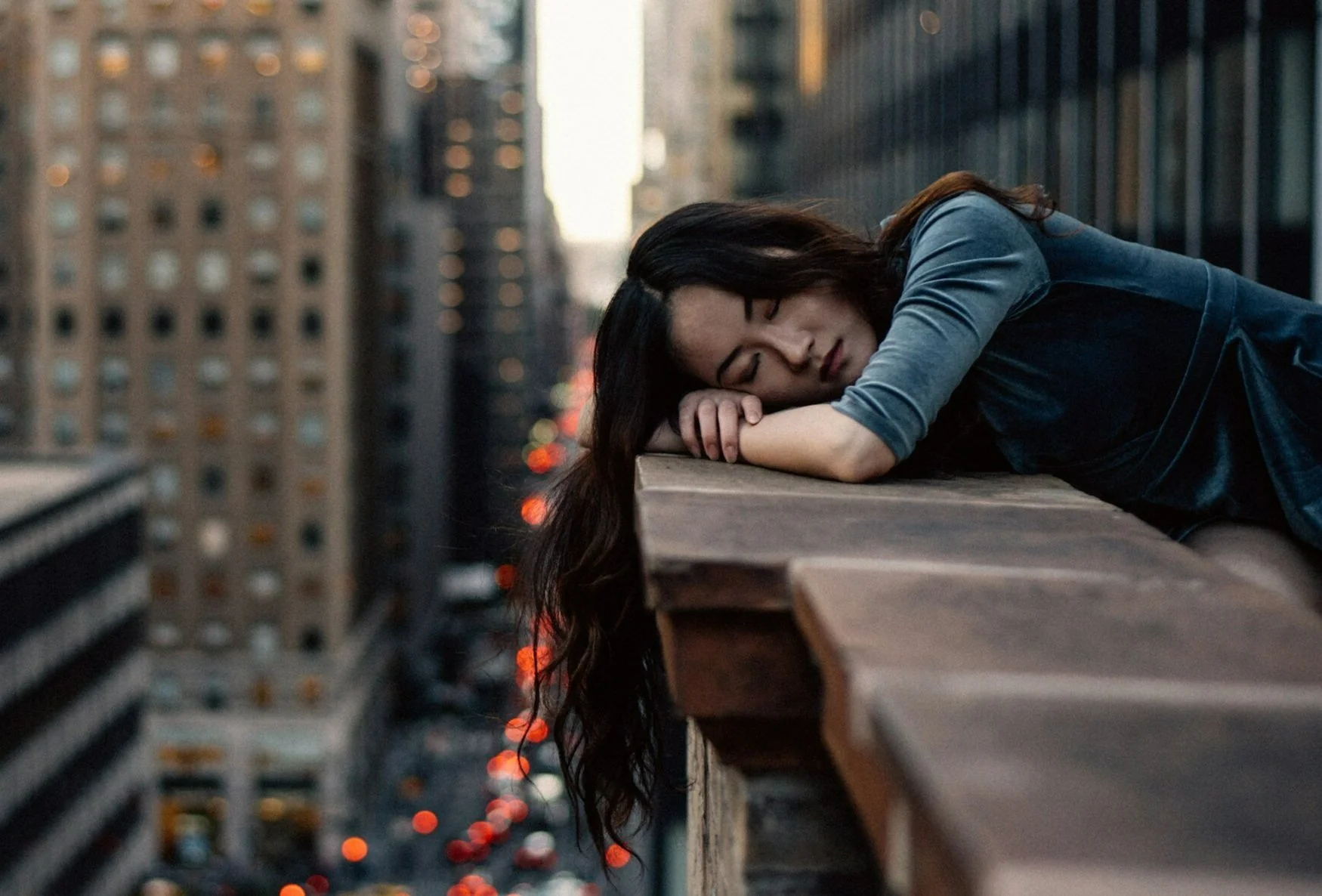 A woman with long dark hair lying on a rooftop railing with her eyes closed, in an urban cityscape at dusk or dawn.