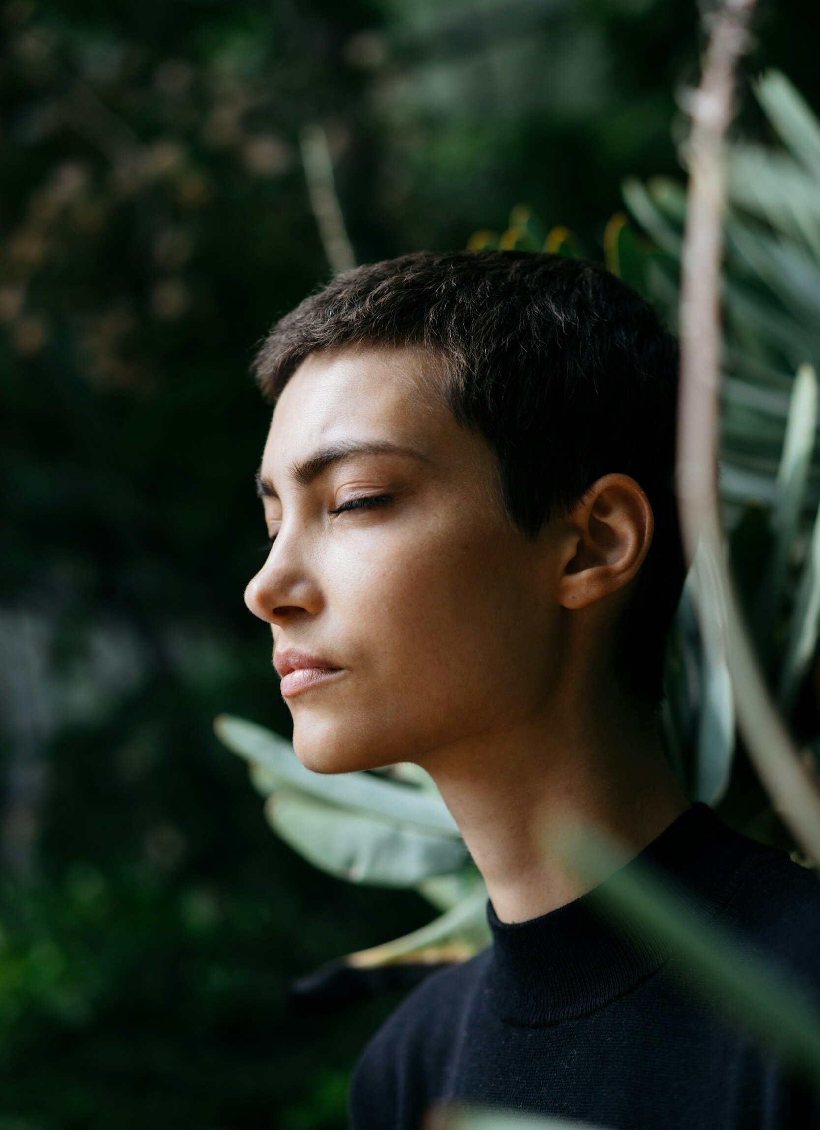 A woman with short dark hair and closed eyes standing in front of lush green foliage.