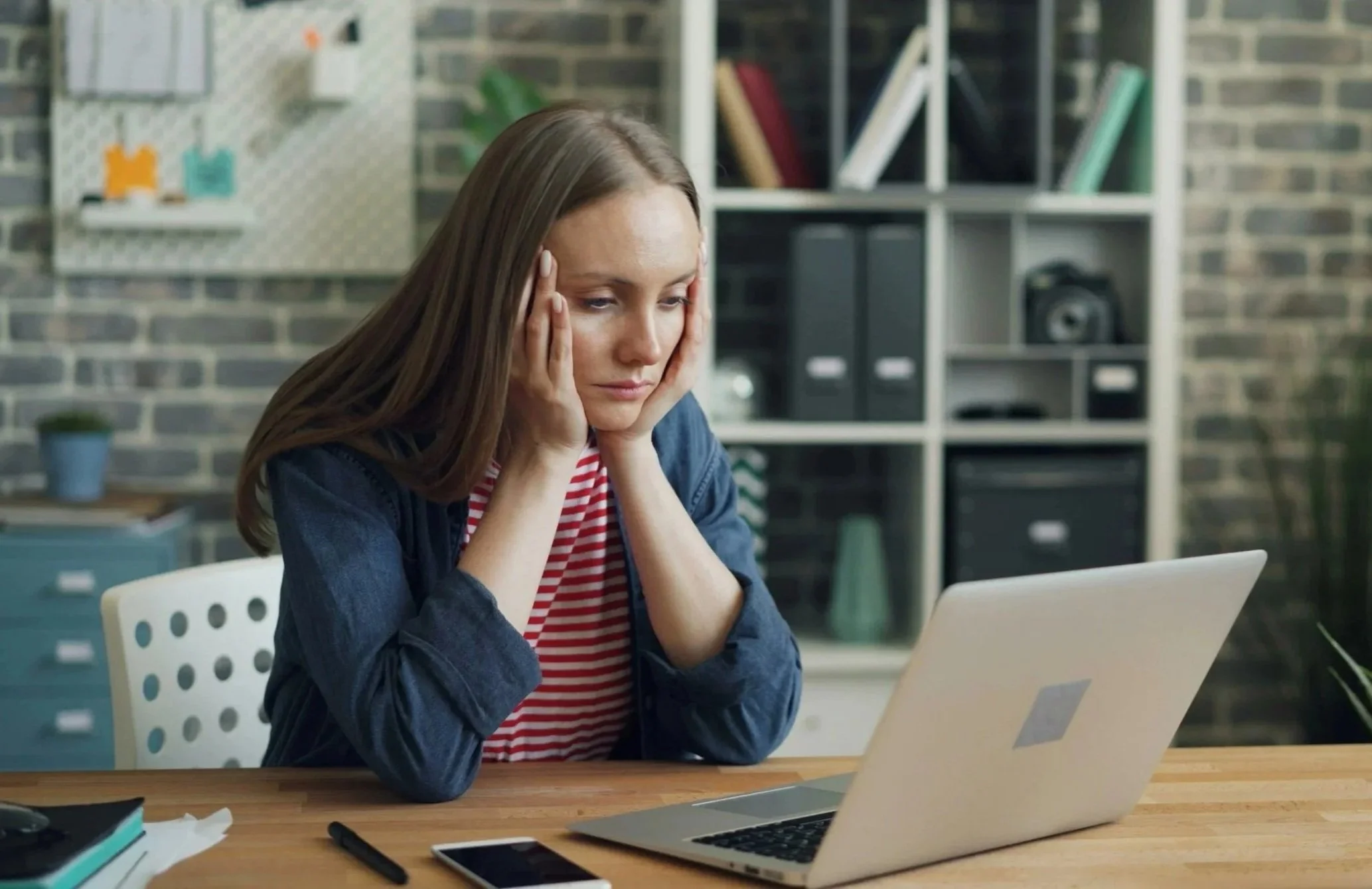 A woman sitting at a desk with her hands on her head, looking stressed while working on a laptop in a modern office.
