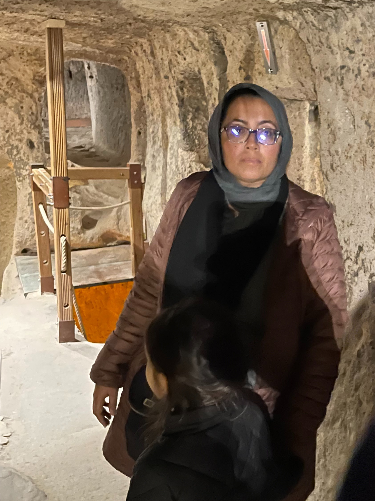 Muslim woman standing in a cave wearing a brown coat and glasses. She is holding her child's hand.