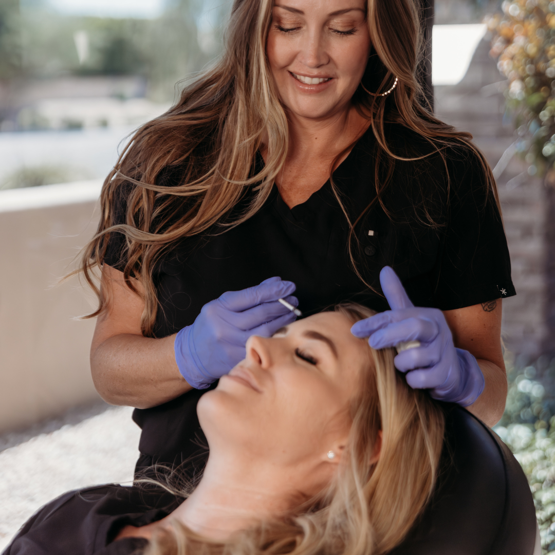 A woman is receiving a cosmetic injection while lying down, smiling, and a professional wearing purple gloves performs the procedure.
