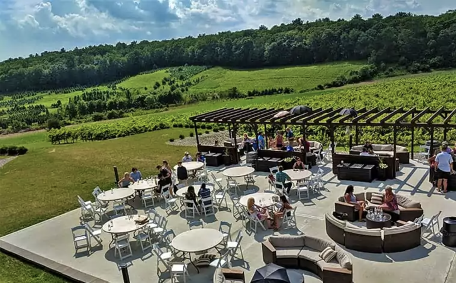 Outdoor patio with tables and chairs, people dining and socializing, surrounded by green fields and rolling hills, under a partly cloudy sky.