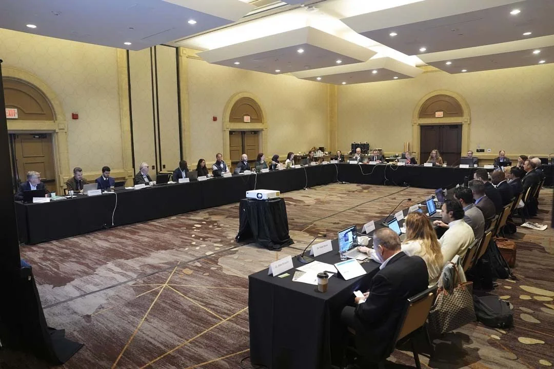 A large conference room with people seated around a U-shaped table, many with laptops, in a formal meeting or discussion setup.