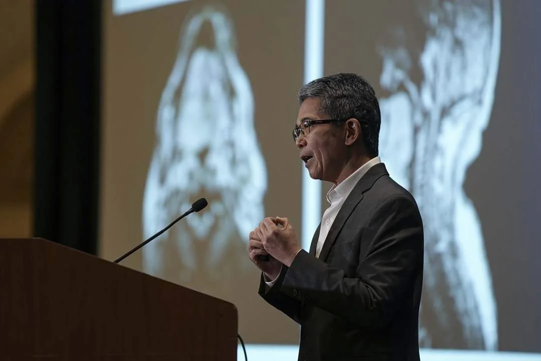 A man giving a presentation in front of a large brain scan image projected on a screen.