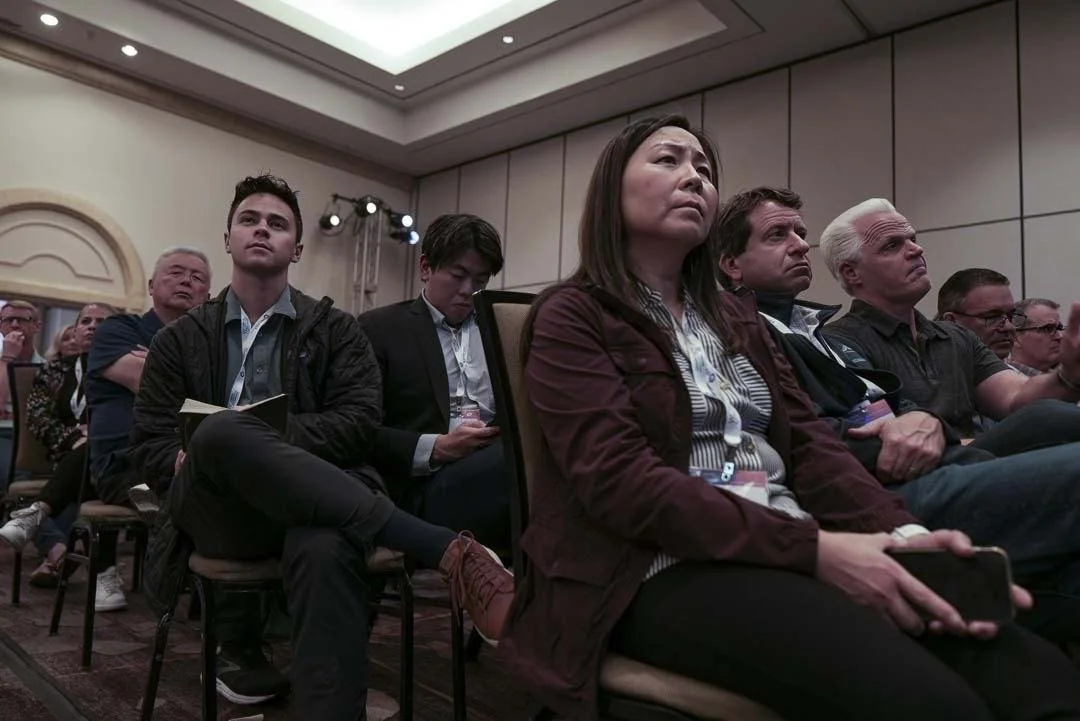 Audience seated in a conference room, attentively listening to a presentation or speaker, with serious expressions.
