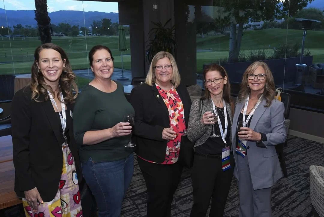 Five women standing indoors at a social event, holding wine glasses, with a view of a golf course visible through large glass windows behind them.