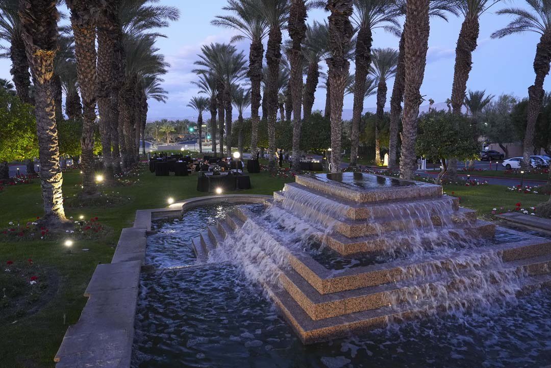 Nighttime outdoor scene with a tiered fountain, surrounded by palm trees, tables with black tablecloths, and string lights, possibly at a garden or event venue.