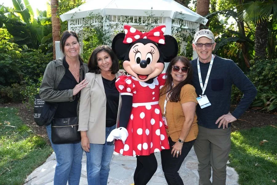 Four adults and a Minnie Mouse mascot posing outdoors in a garden area with trees and lush green plants.