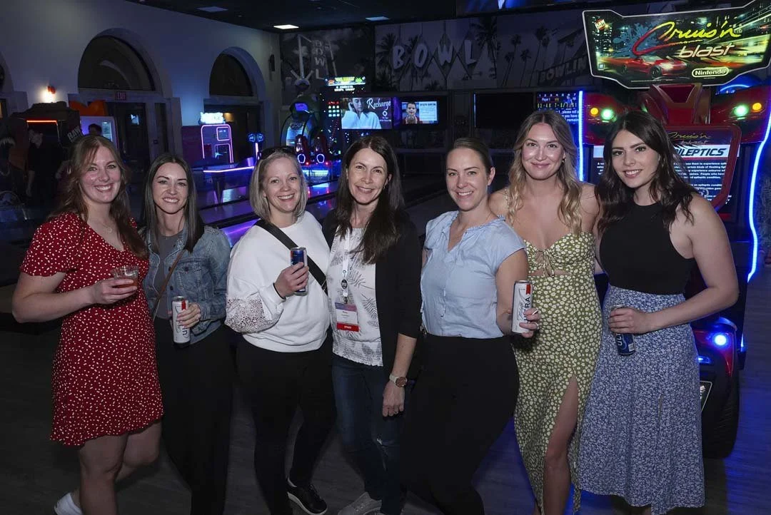 A group of seven women smiling together at an arcade, with bowling lanes and arcade games in the background.