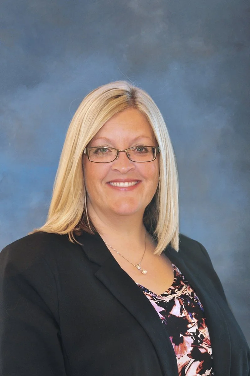 A professional woman with blonde hair, wearing glasses, a black blazer, and a patterned blouse, smiling in front of a blue-gray background.