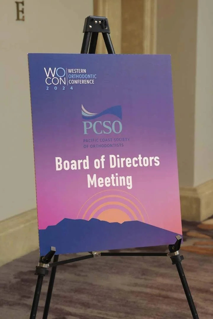A sign on an easel advertising the Board of Directors Meeting for the Pacific Coast Society of Orthodontists, part of the Western Orthodontic Conference 2024, with a sunset over mountains in a purple and pink gradient background.