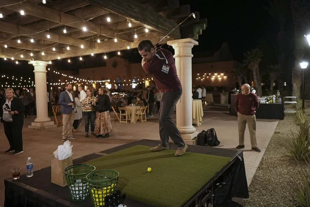 A man in a maroon shirt and dark pants swings a golf club at a yellow golf ball on a small platform at an outdoor event under string lights at night.